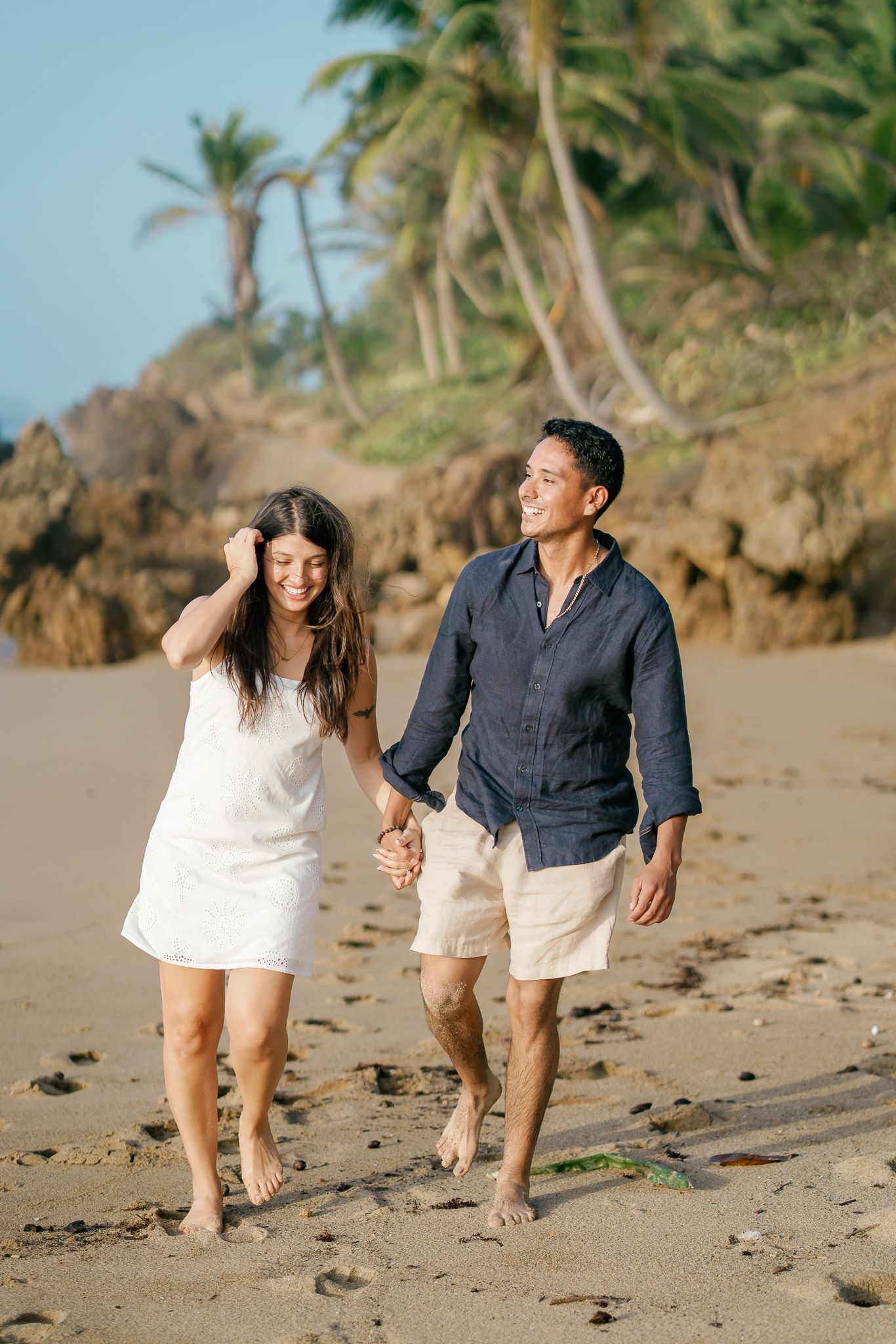 A couple in white and navy casual attire walks playfully together along a tropical beach with palm trees in the background.