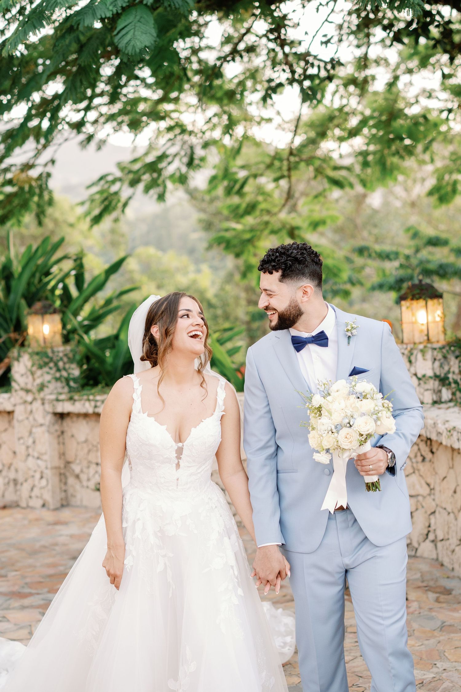 Bride and groom holding hands laughing outdoors, she in lace gown with veil, he in light blue suit with bow tie holding bouquet.