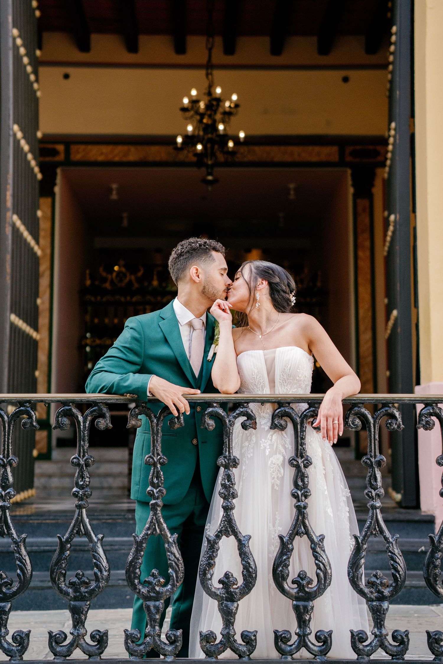 Bride and groom share a romantic kiss on an ornate balcony, groom in green suit, bride in strapless lace gown.
