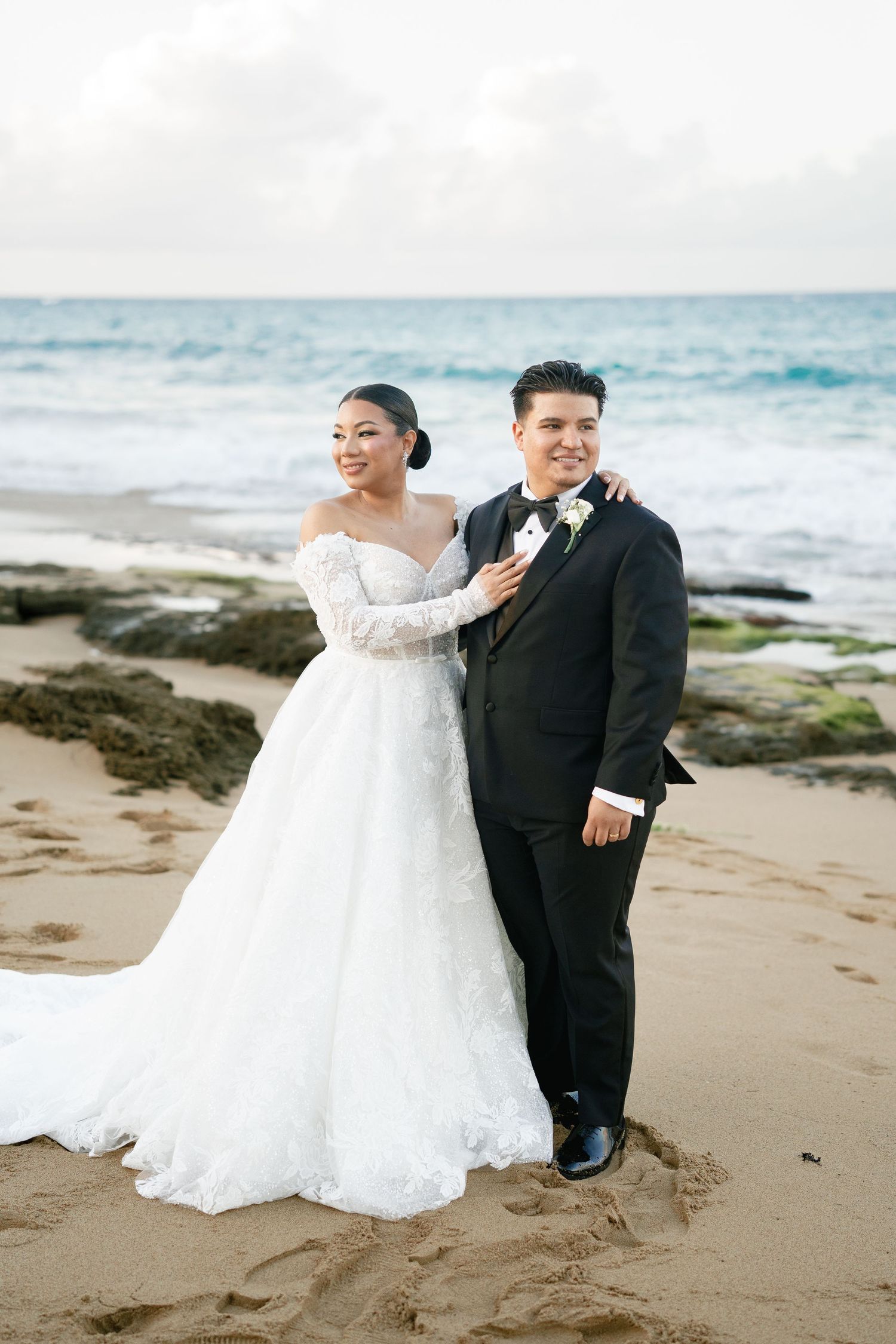 Bride in lace off-shoulder gown and groom in black tuxedo pose together on sandy beach with ocean backdrop.