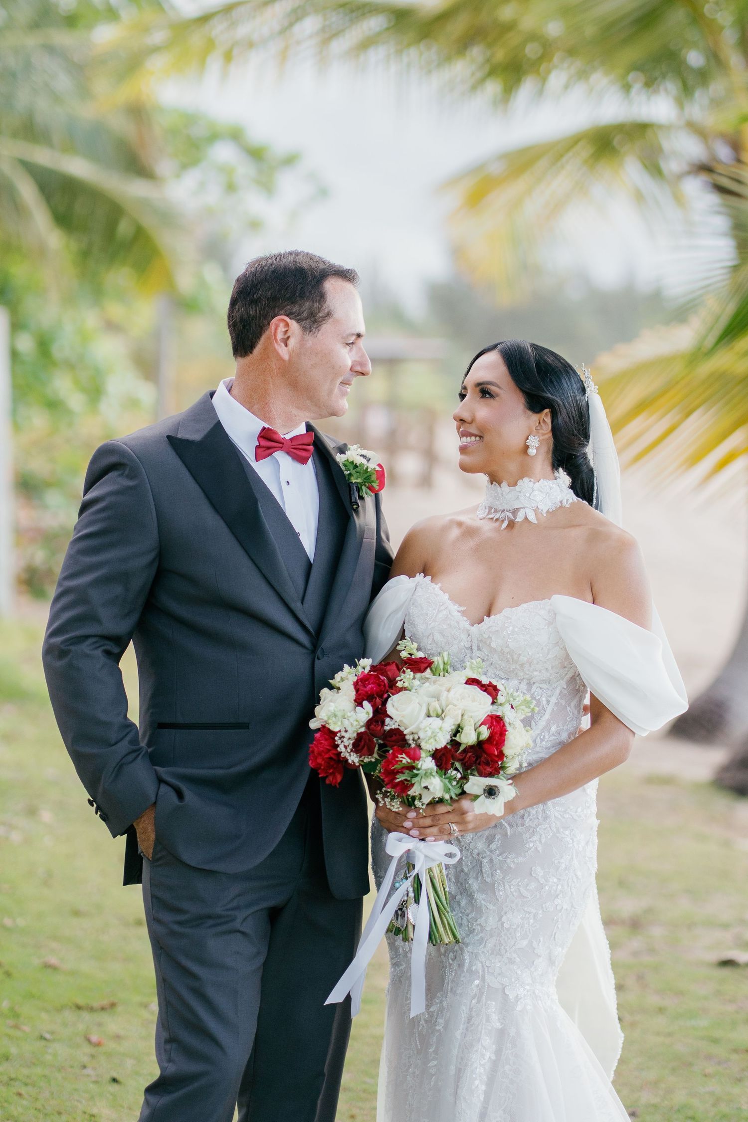Bride and groom share a romantic gaze outdoors, she holds red and white bouquet, wearing off-shoulder gown, he in dark tuxedo.