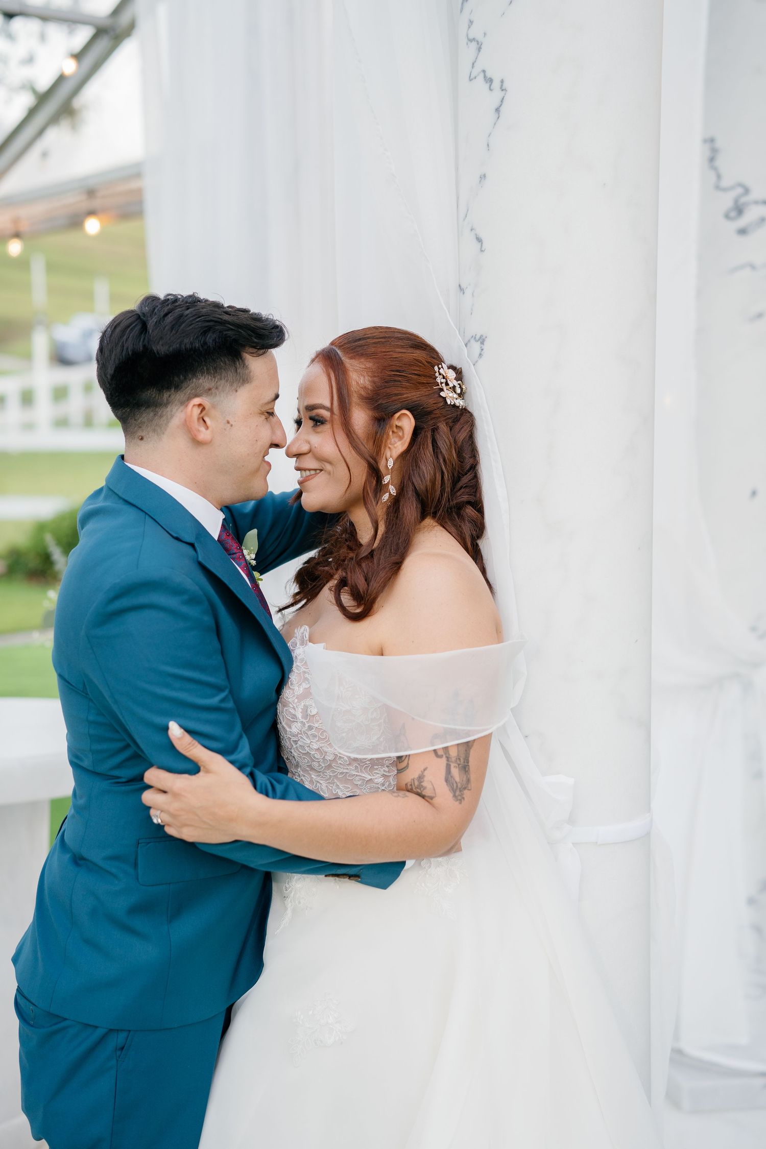 A bride and groom share a romantic embrace near marble columns, smiling at each other on their wedding day.