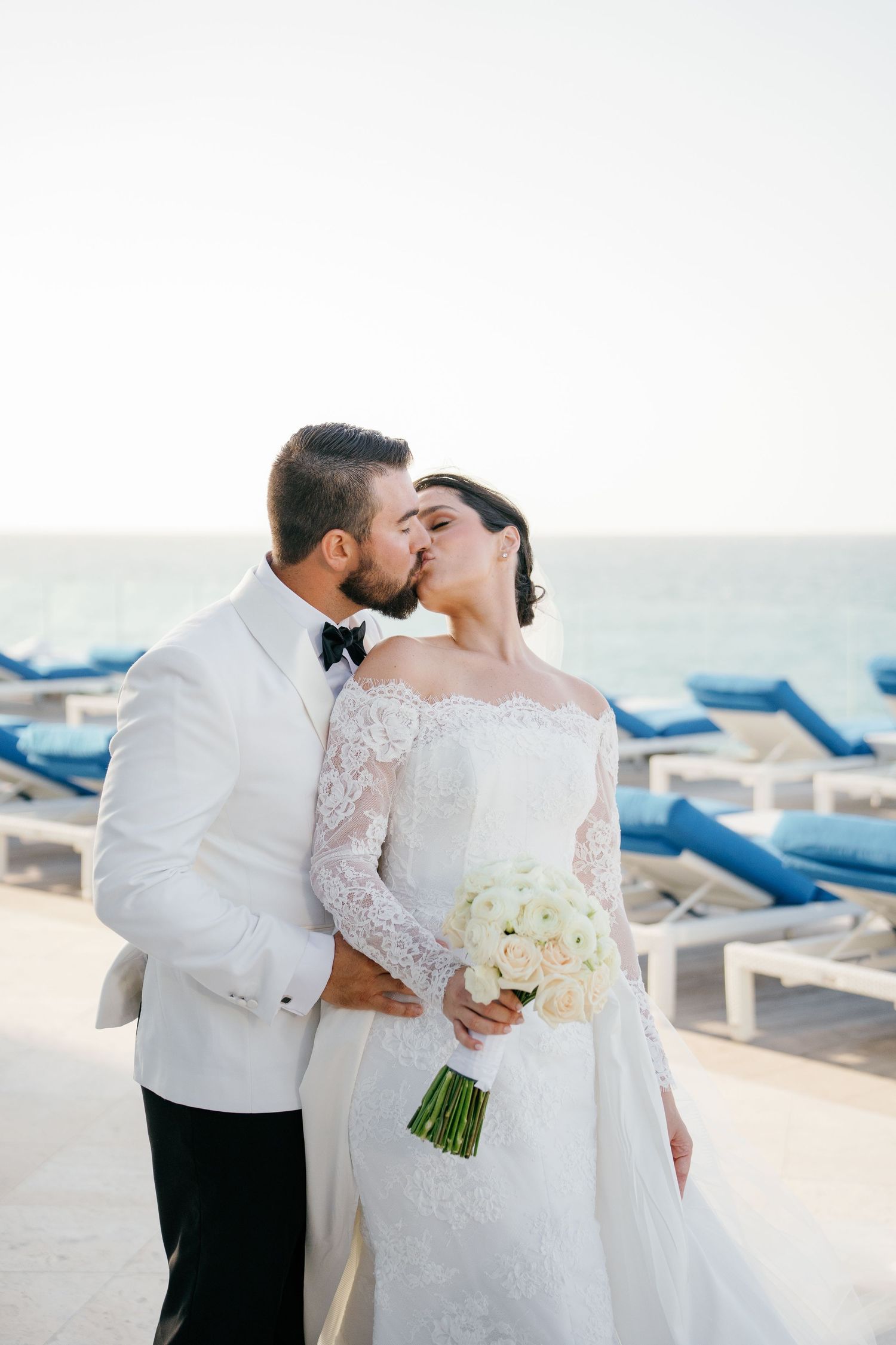 Bride and groom share a romantic kiss at a beachside venue, bride holding white bouquet in lace wedding gown.