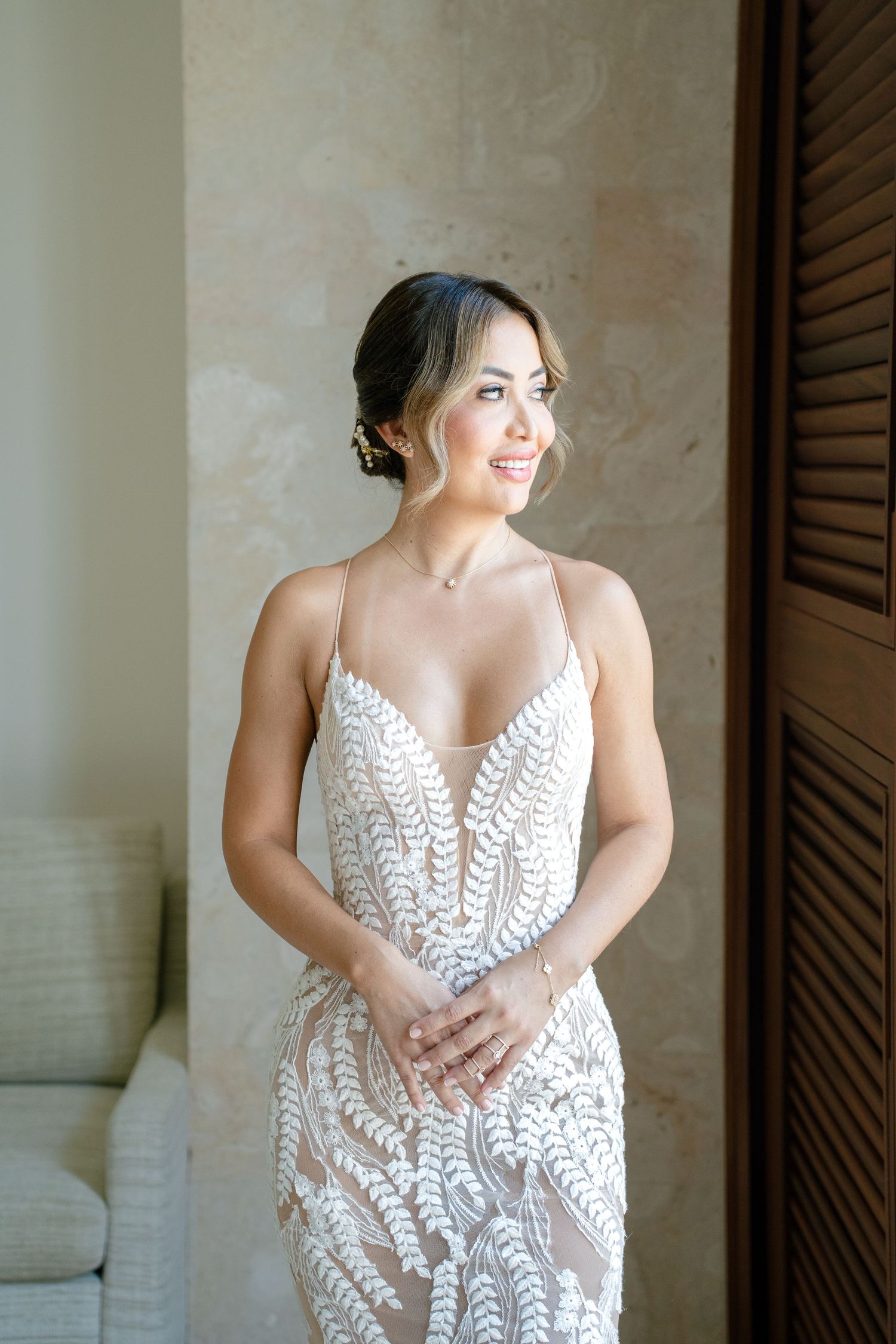 Bride in elegant white lace gown posing in studio against textured wall with wooden shutters.