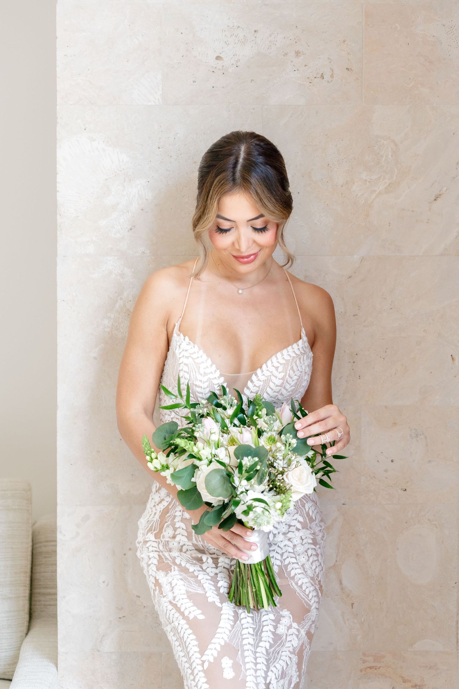 Elegant bridal portrait featuring a lace dress and eucalyptus bouquet against a neutral wall.
