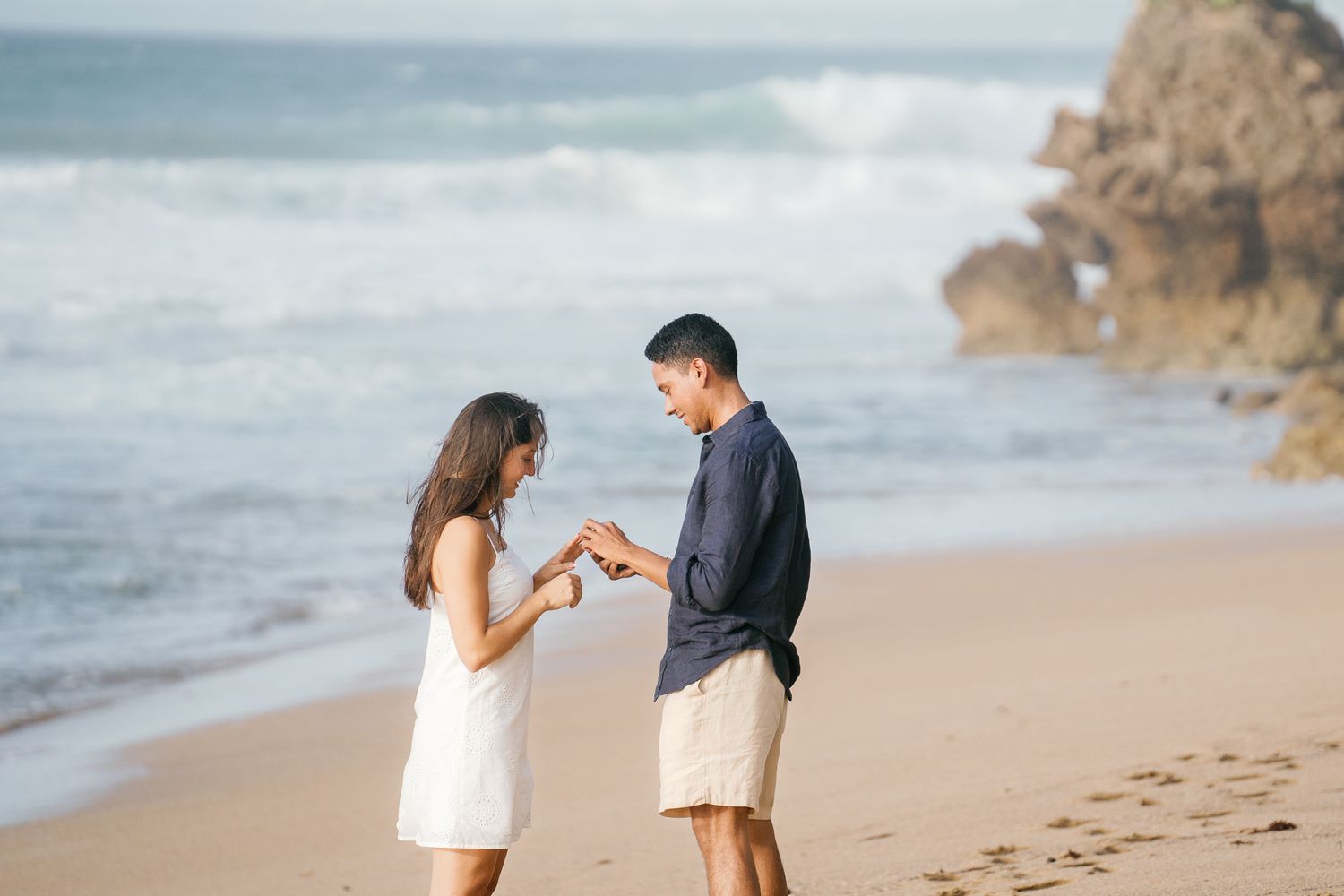 Two people walking together on a sandy beach with ocean waves and rocky cliffs visible in the distance.