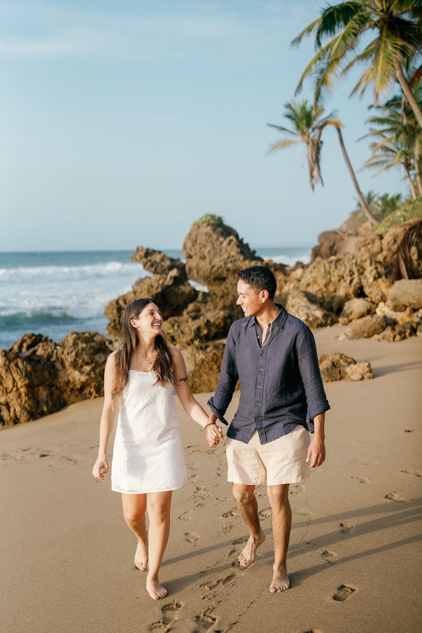 A couple walks hand in hand along a tropical beach with rocky outcrops and palm trees in the background.