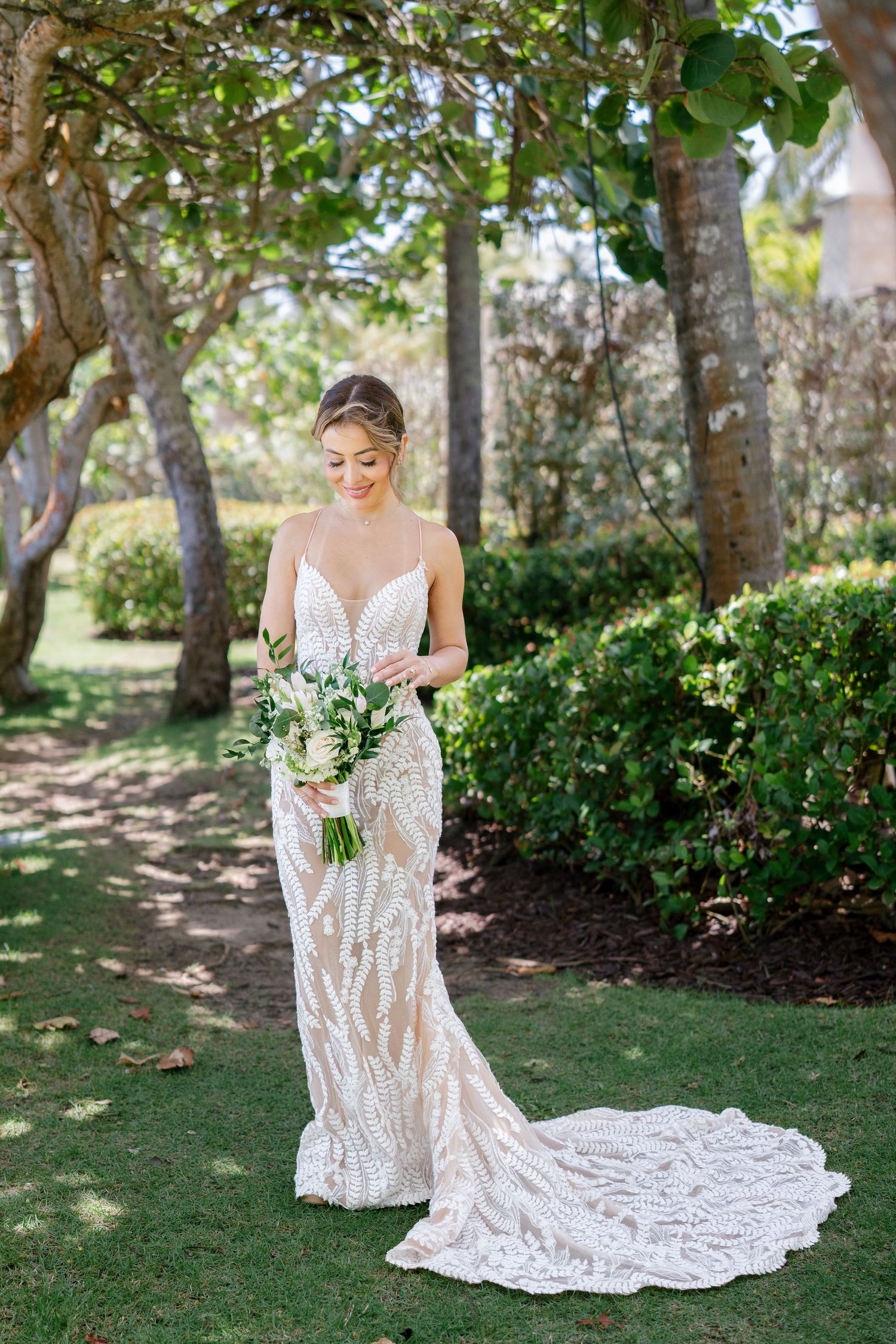 A bride in a flowing lace wedding gown with train holds a green and white bouquet in a garden setting.
