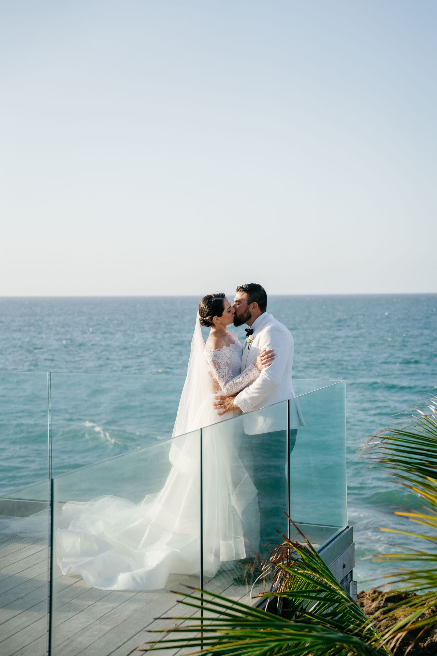 Bride and groom share a romantic kiss on an oceanfront balcony, bride wearing a flowing white gown and veil.