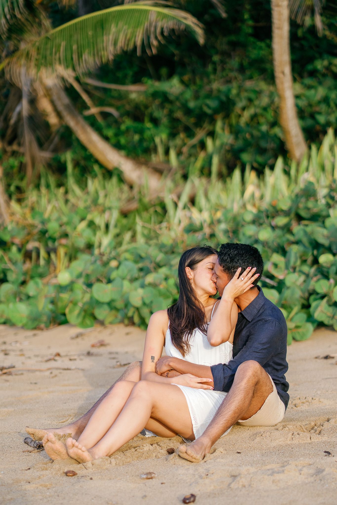 A couple embraces while relaxing together on a sandy beach with tropical foliage in the background.