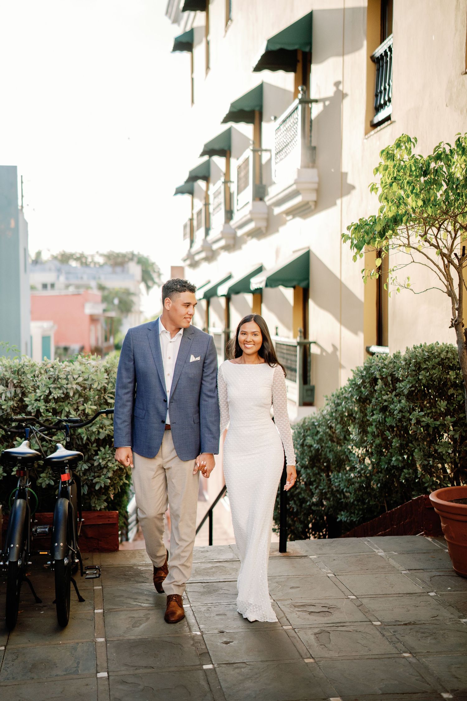 Couple walking hand in hand on a sunny sidewalk, man in blue blazer, woman in elegant white gown, beside a classic building.