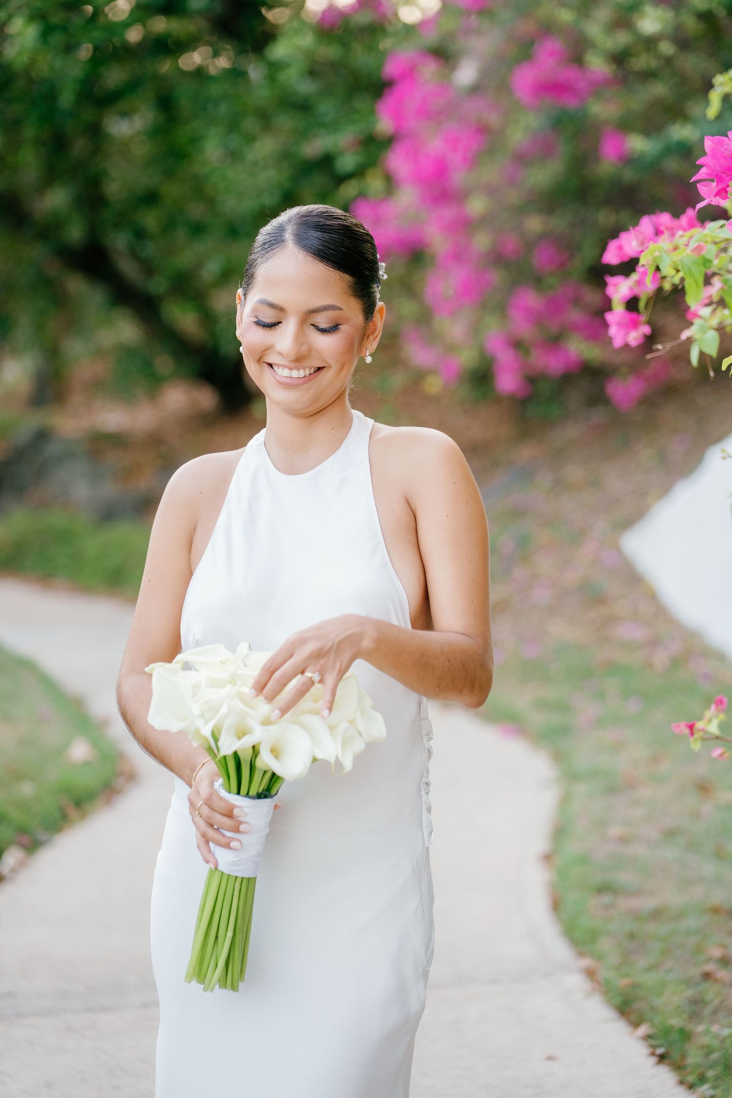 Smiling bride in white halter gown holding calla lily bouquet outdoors with pink bougainvillea blooms in background.