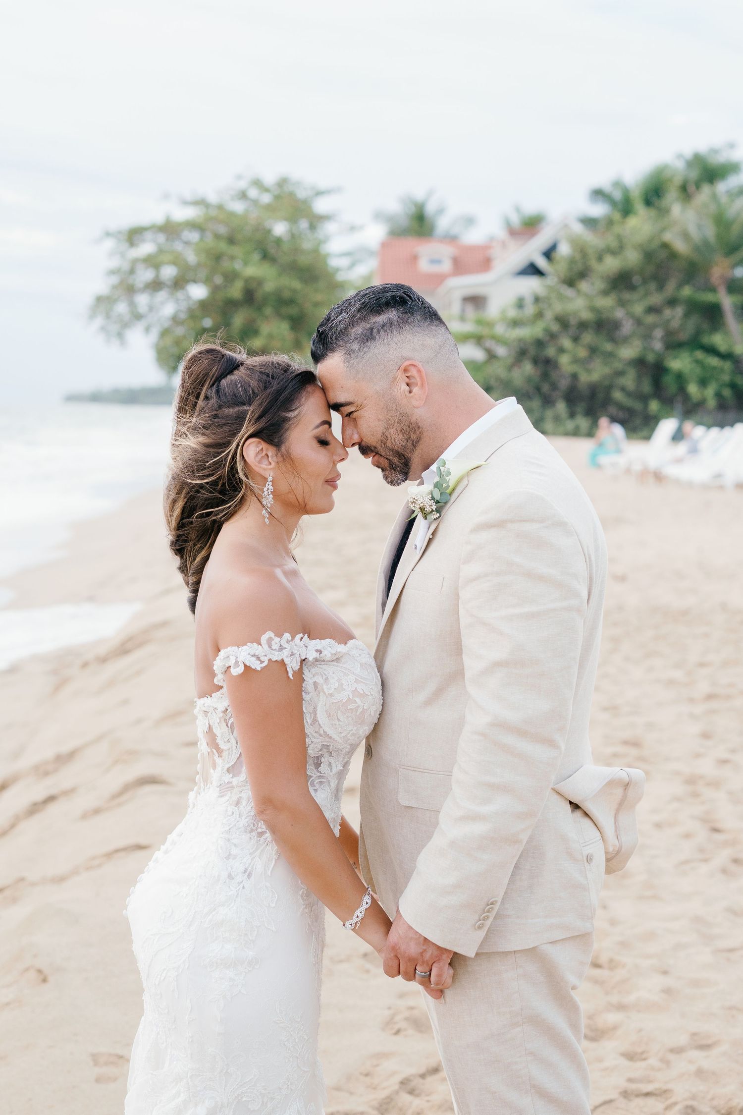 Bride and groom holding hands on a sandy beach, touching foreheads romantically, wearing lace gown and beige suit.
