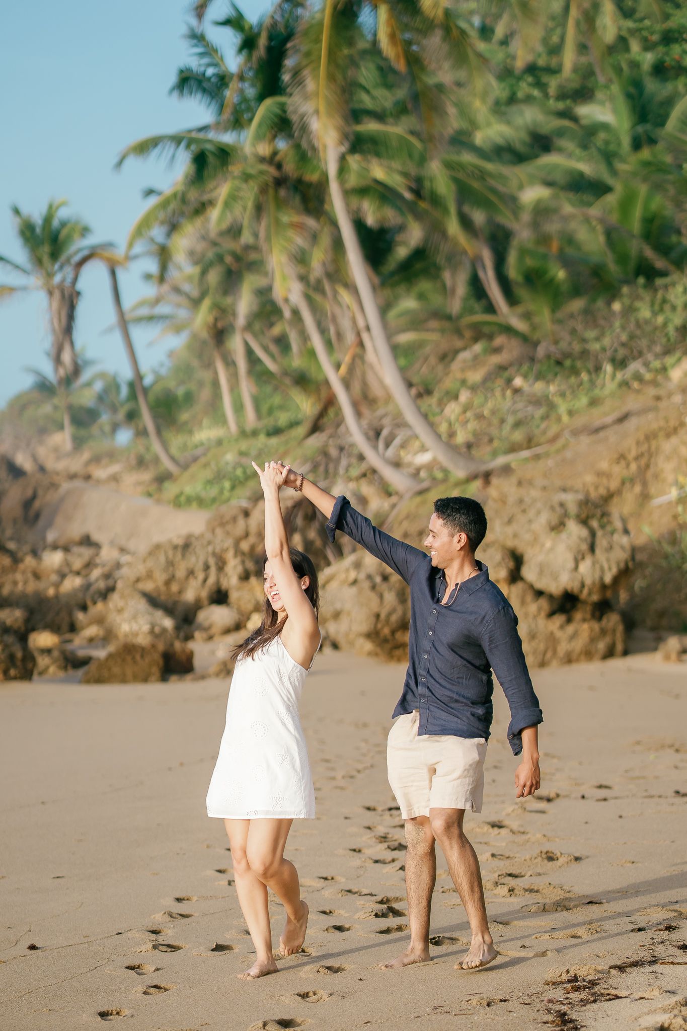 A couple dances playfully together on a tropical beach with palm trees swaying in the background.