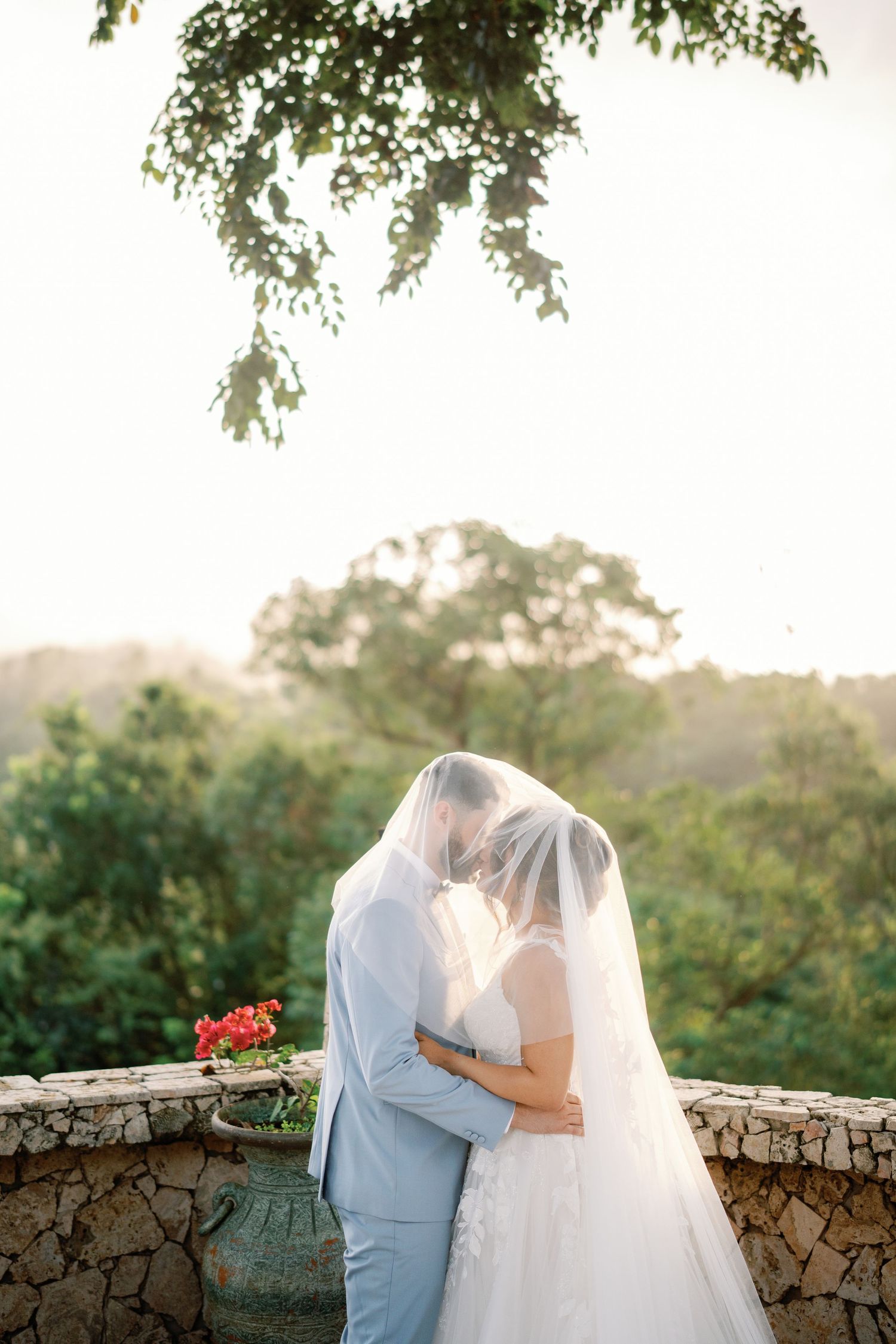 A bride and groom share a romantic kiss under a flowing veil at an outdoor wedding with lush greenery backdrop.
