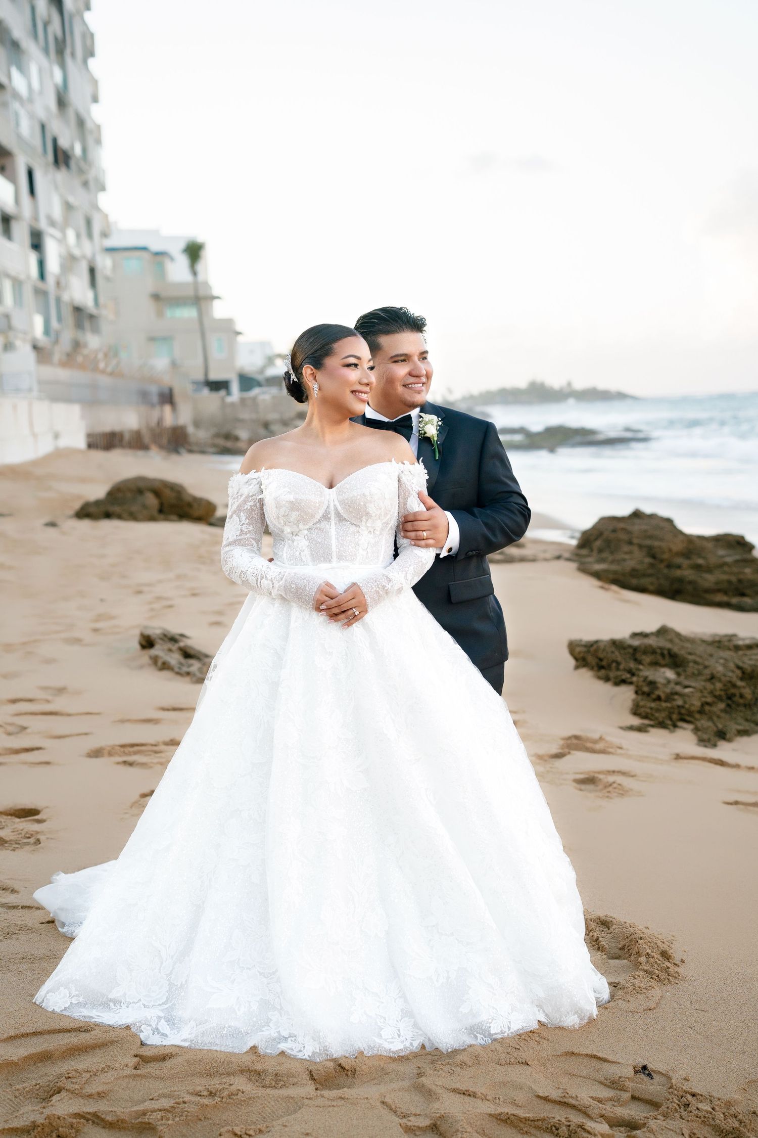 Bride in off-shoulder lace ball gown and groom in navy suit pose romantically on sandy beach with ocean backdrop.