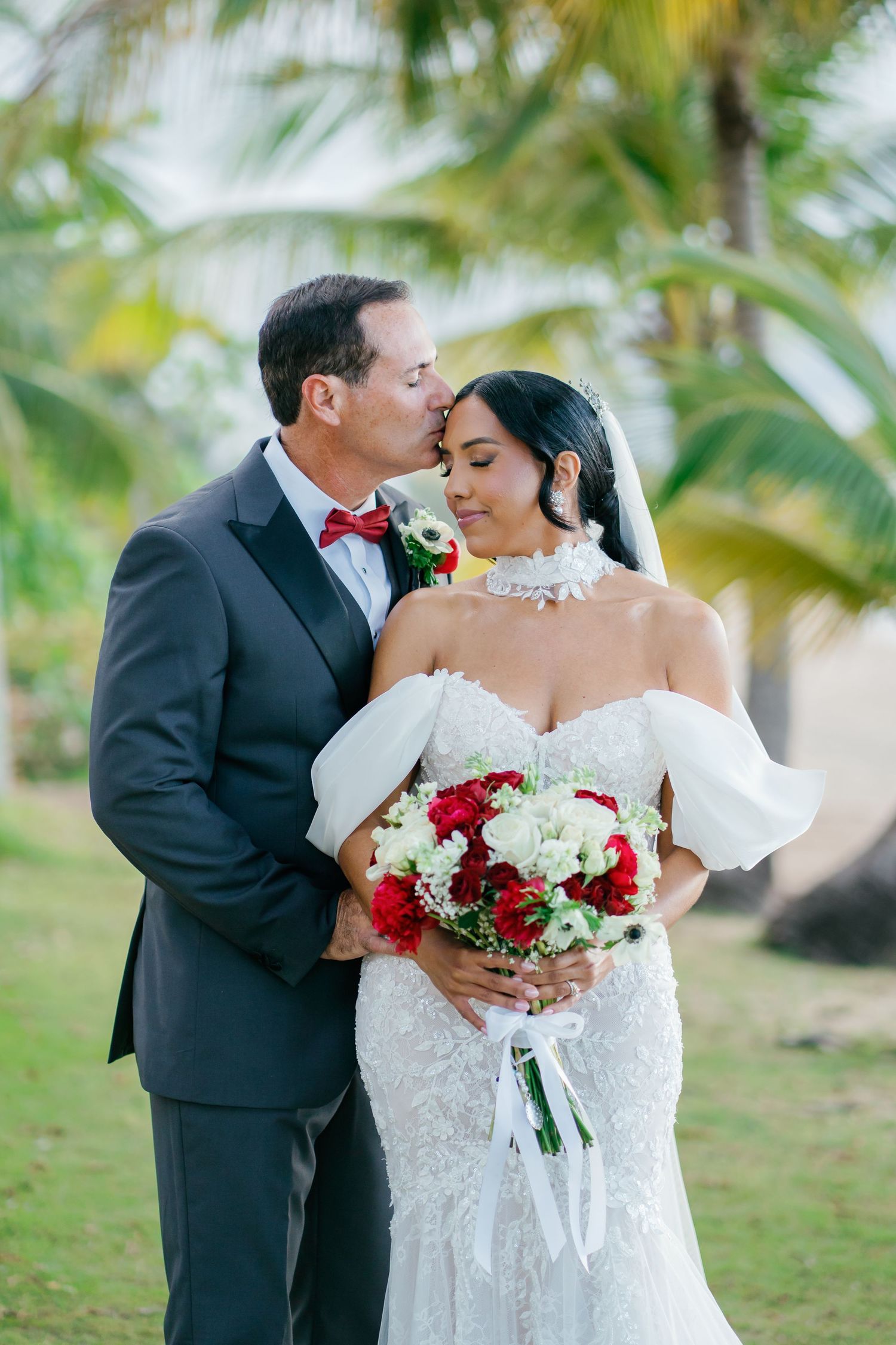 Groom in dark suit kissing bride in lace off-shoulder gown holding red and white bouquet outdoors.