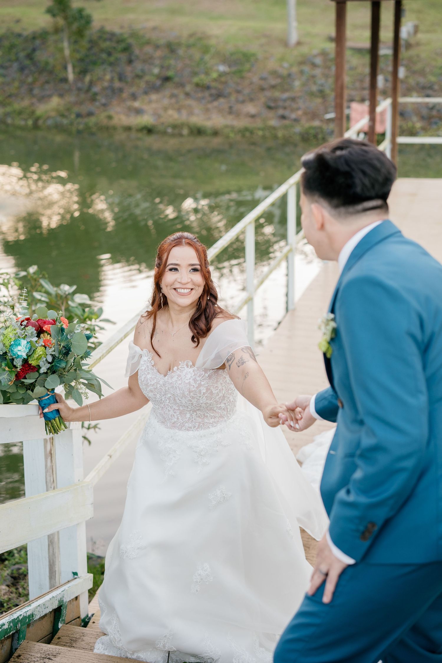 Bride in white gown holds groom's hand on lakeside dock, smiling while holding colorful bouquet.