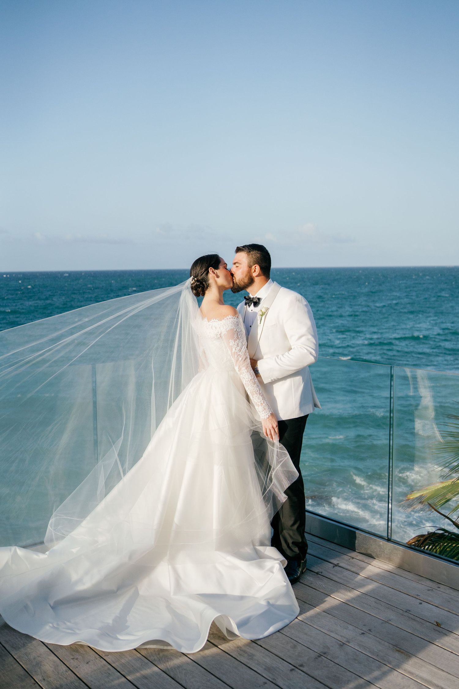 Bride and groom share a romantic kiss on an oceanfront deck, her flowing veil billowing in the breeze.