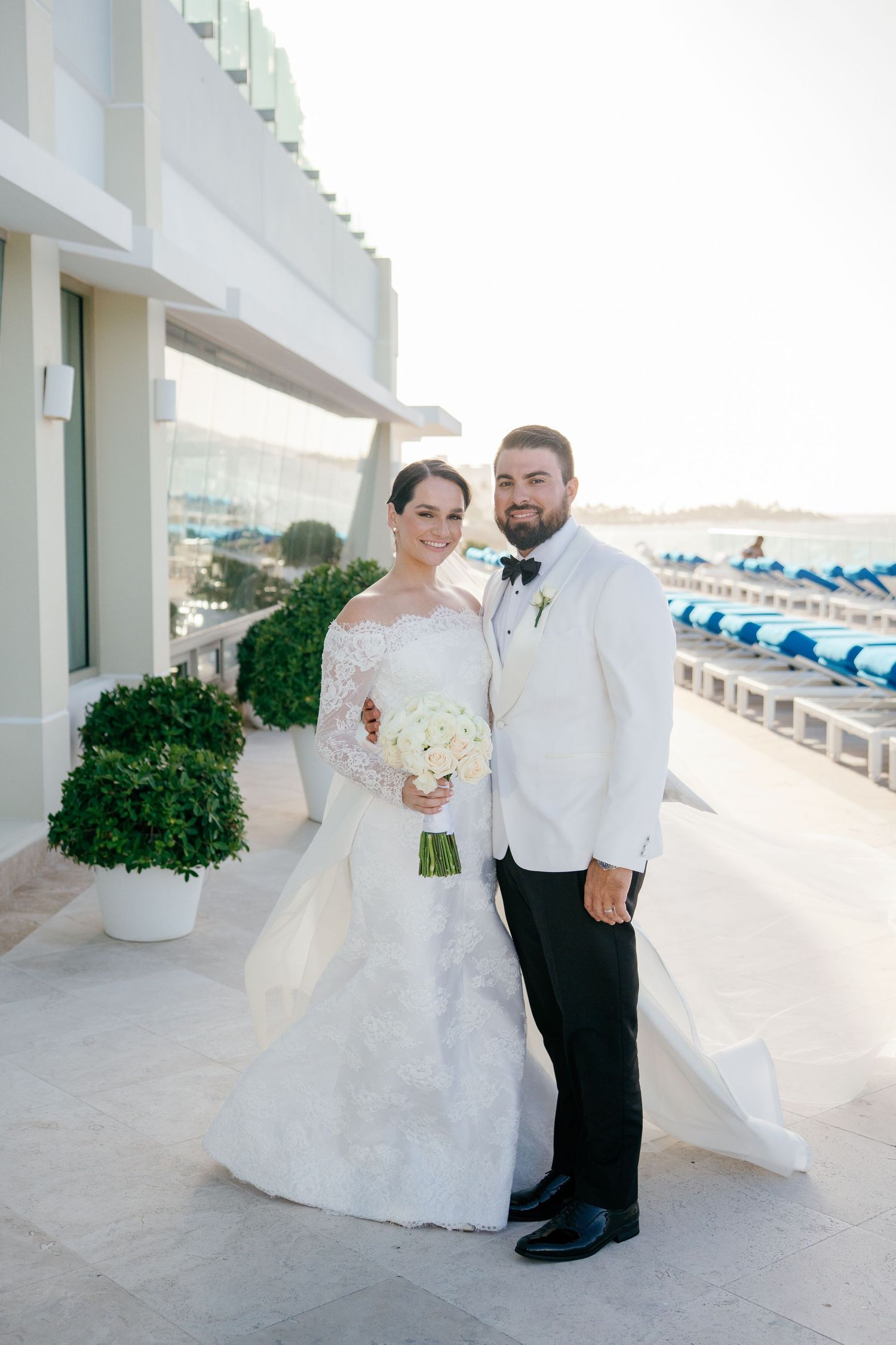 Bride and groom pose together on a waterfront terrace, she in lace gown holding bouquet, he in white tuxedo jacket.