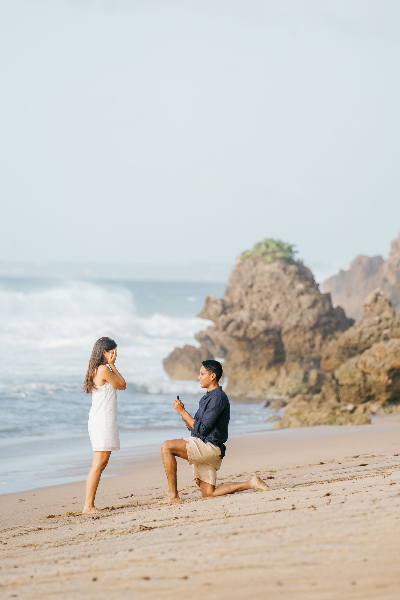 A romantic beach proposal scene with rocky coastal cliffs and crashing waves in the background.