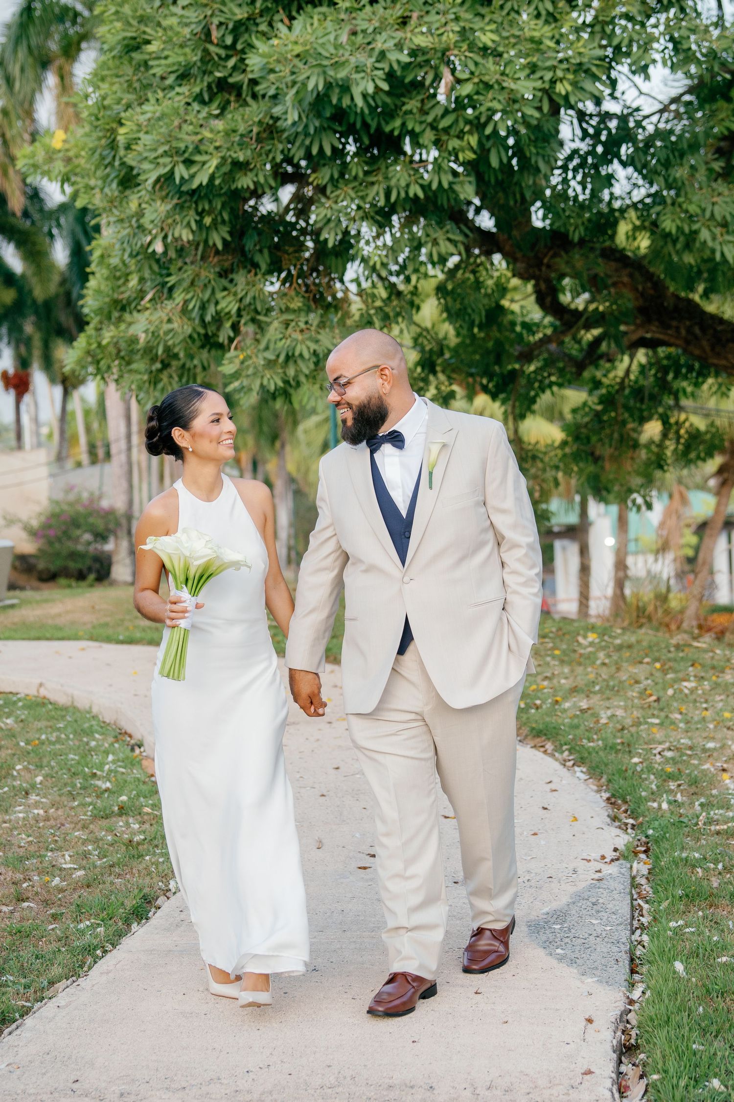 Bride and groom walking hand-in-hand on a garden path, bride in white gown holding bouquet, groom in beige suit.