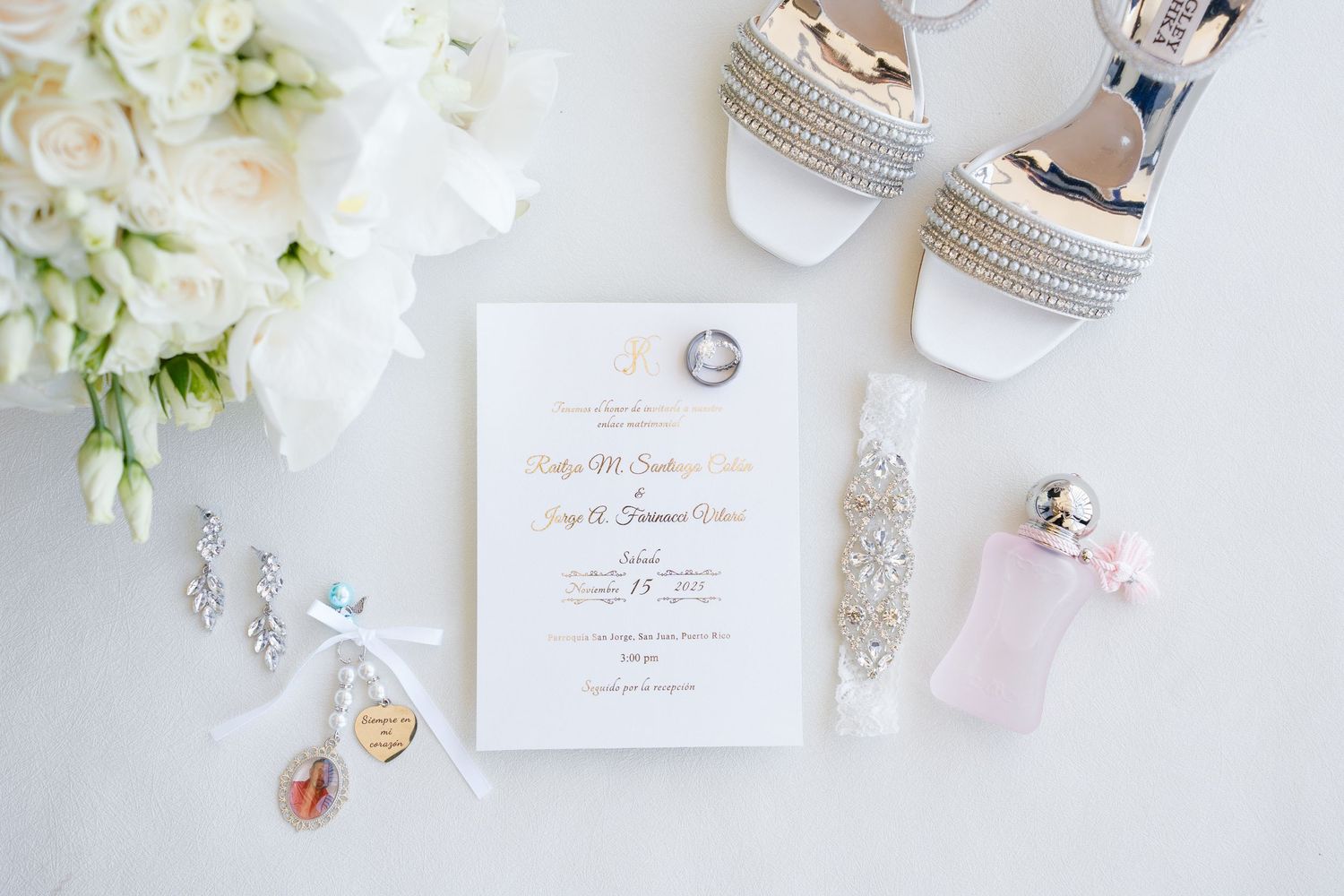 Elegant wedding flat lay featuring invitation suite, jeweled heels, floral bouquet, perfume, garter, and diamond rings on white background.
