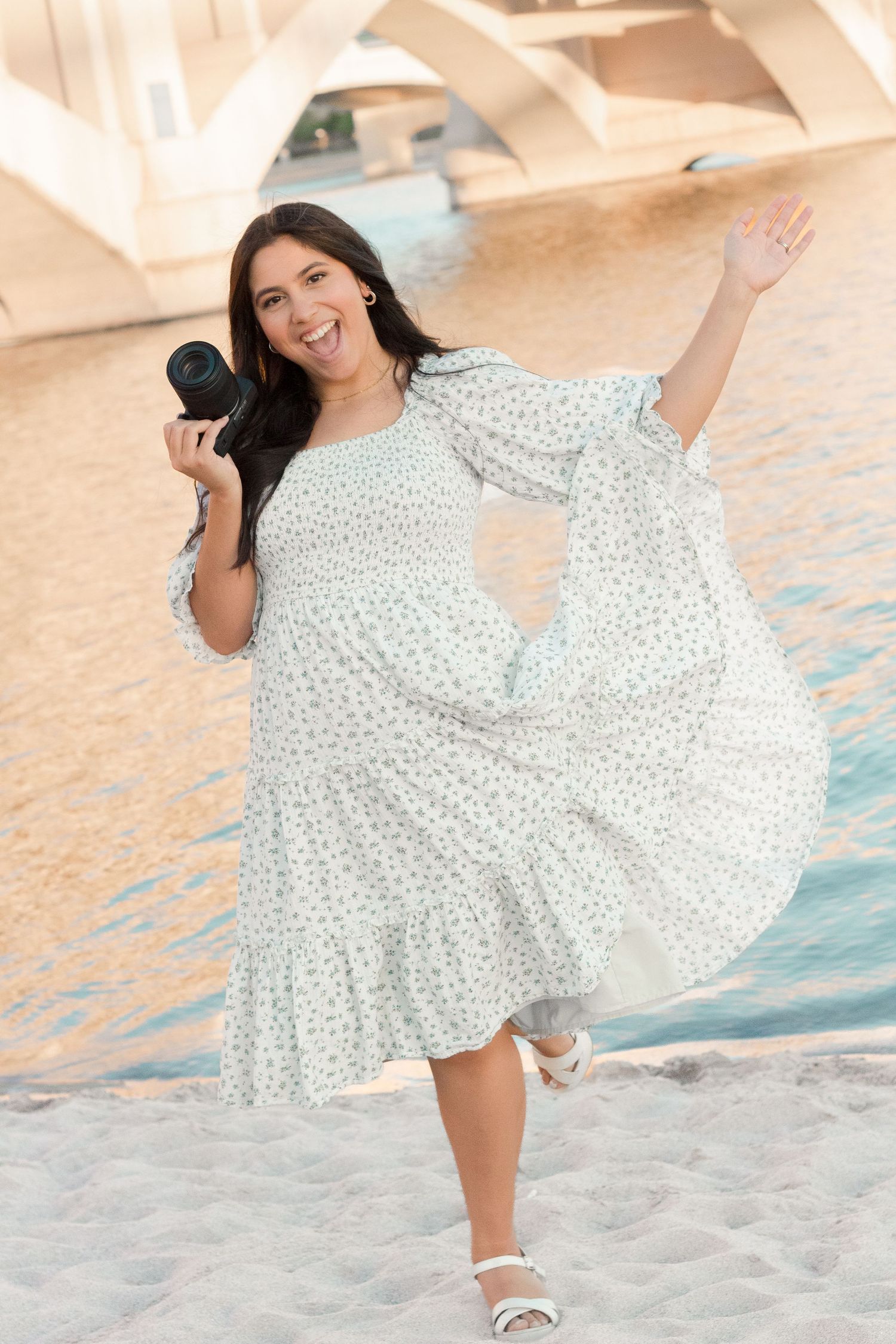 Portrait of a young family photographer girl smiling with camera in hand and throwing dress in the air at Tempe Town Lake.