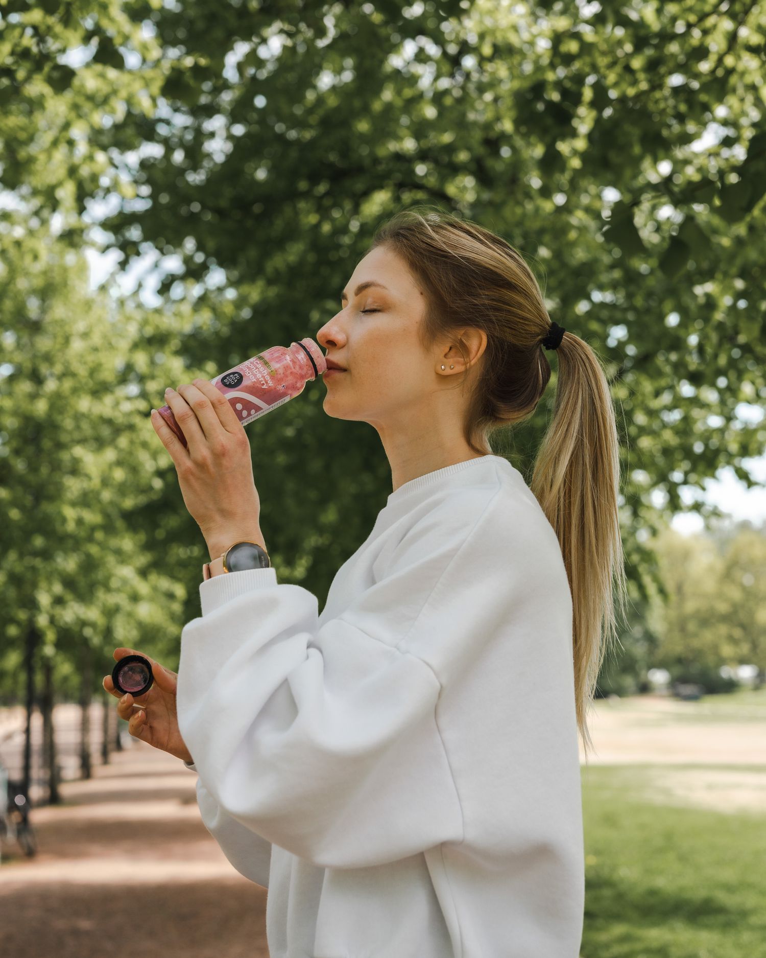 A person in a white sweatshirt drinking from a pink bottle while standing on a tree-lined path on a sunny day.