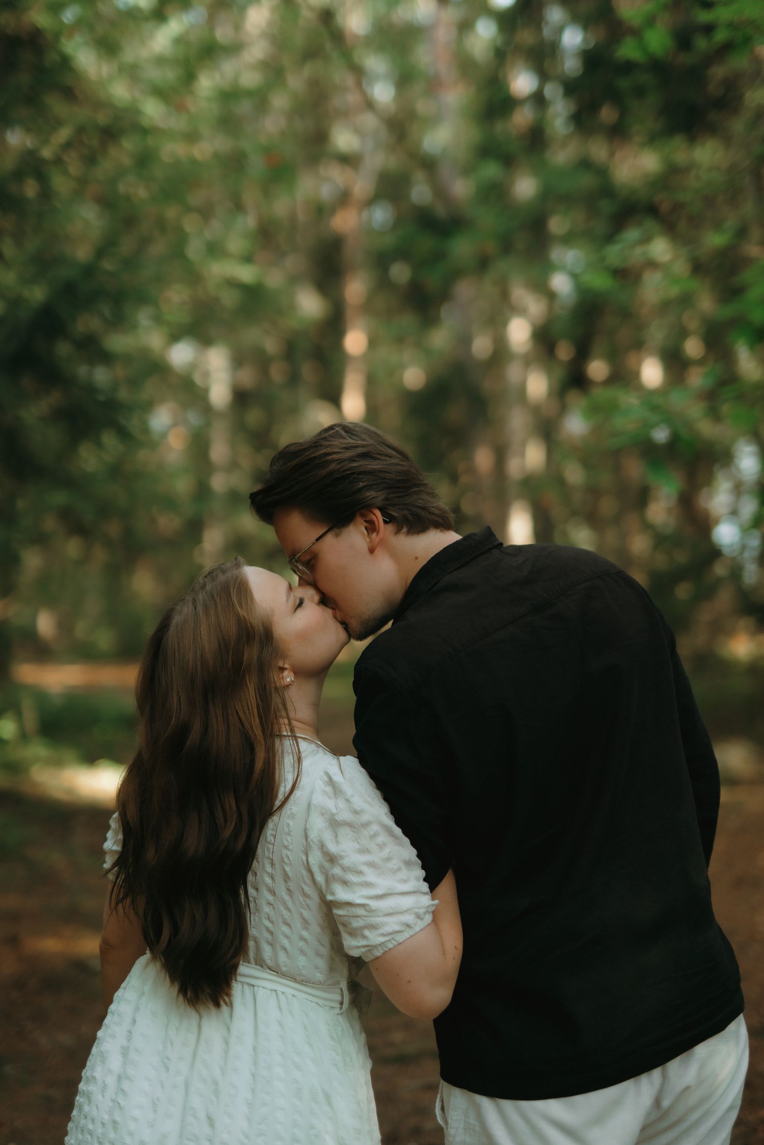 A romantic couple share a tender kiss in a sunlit forest, she in a white dress and he in dark attire.
