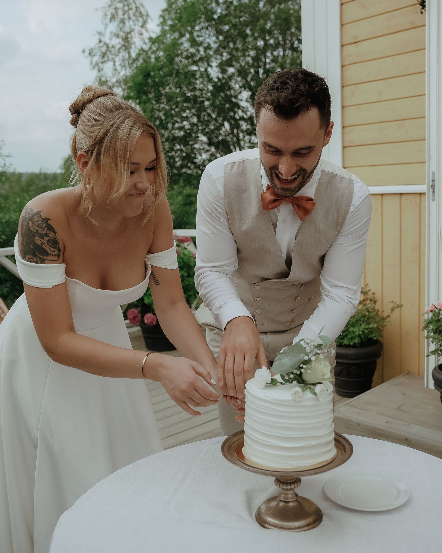 A couple in wedding attire cuts their simple white wedding cake together on a modern gold cake stand outdoors.