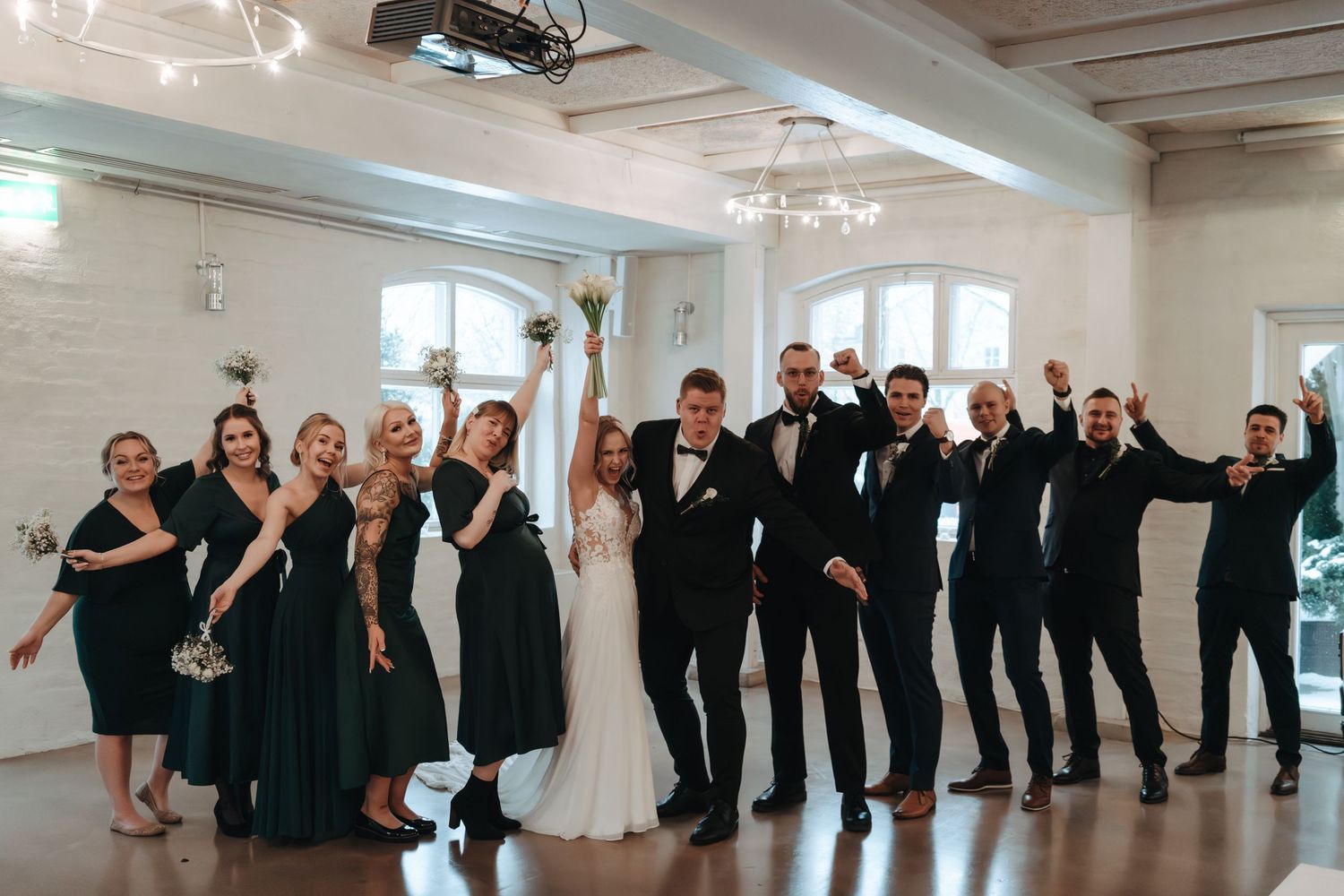 A wedding party in formal black and white attire poses together in a bright indoor venue for a fun group photo.