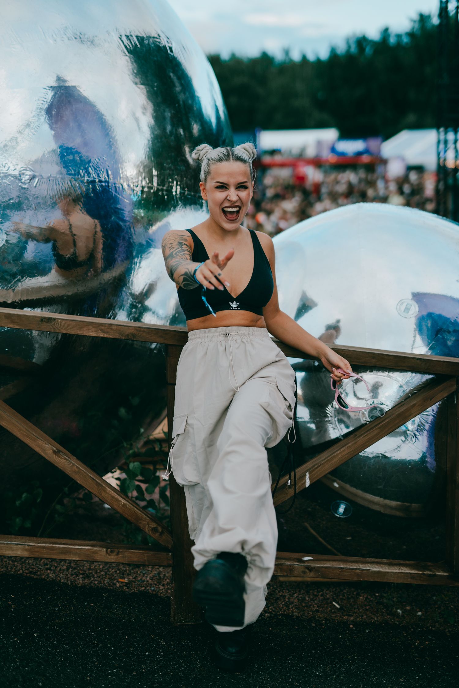 A festival-goer in black crop top and baggy white pants poses against a wooden railing at an outdoor event.