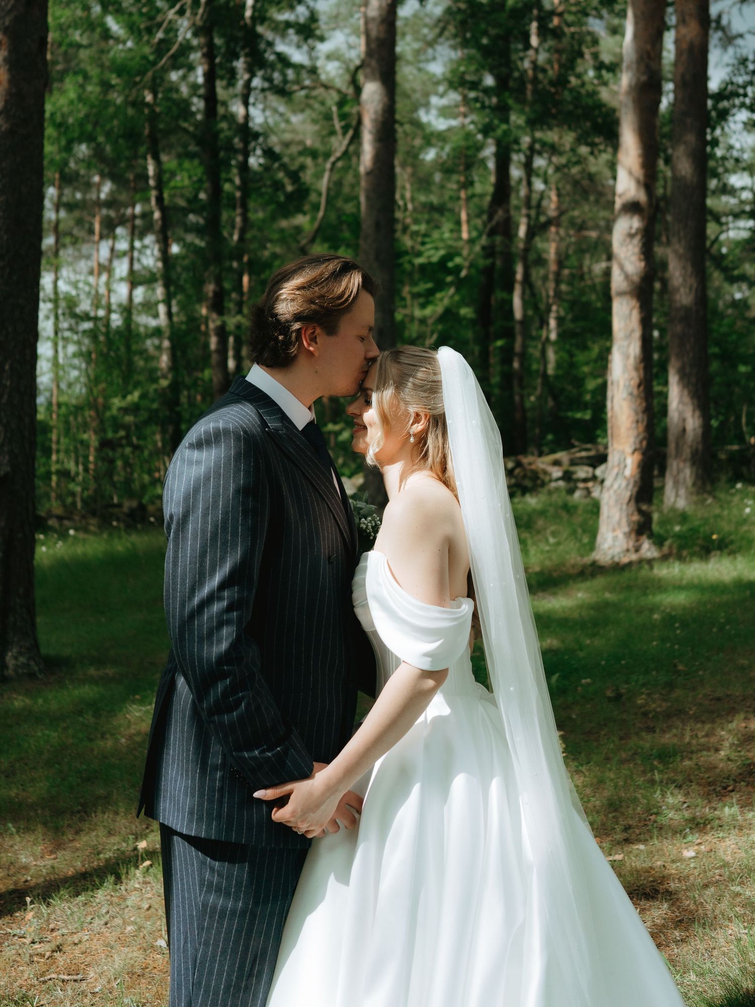 A bride and groom share a kiss in a sunlit forest, she in a white off-shoulder gown with veil, he in a pinstripe suit.