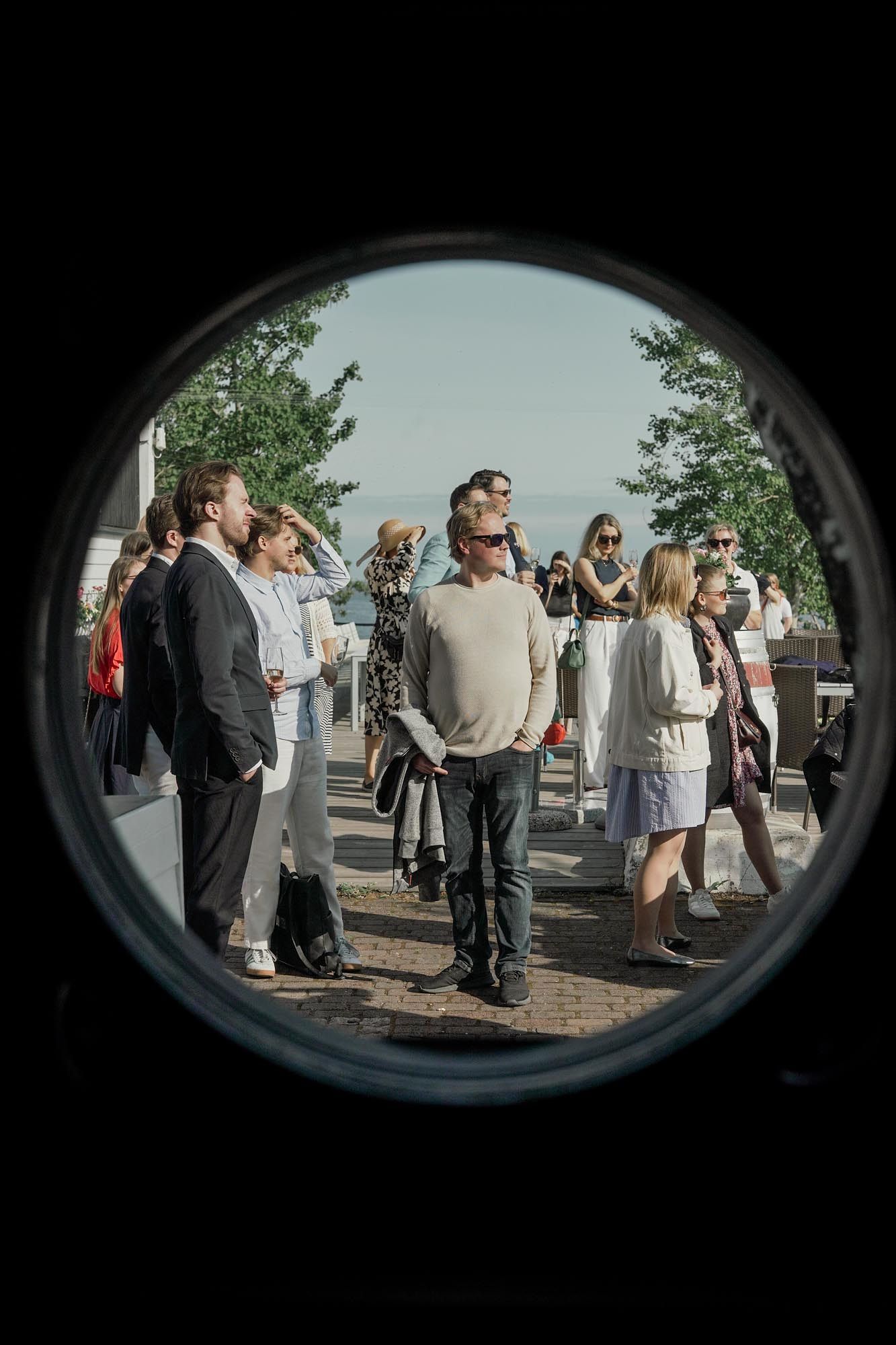 Group of elegantly dressed people gathered outdoors, viewed through a circular porthole frame on a sunny day.