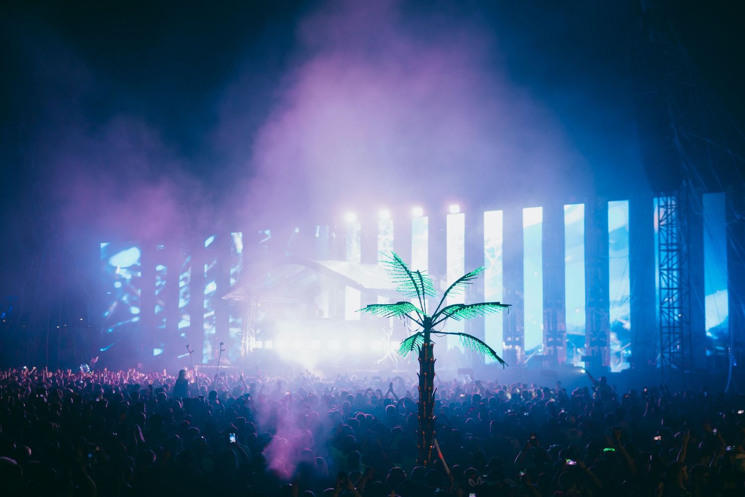 Illuminated palm tree silhouette at a music festival with bright blue lights and fog effects on stage.