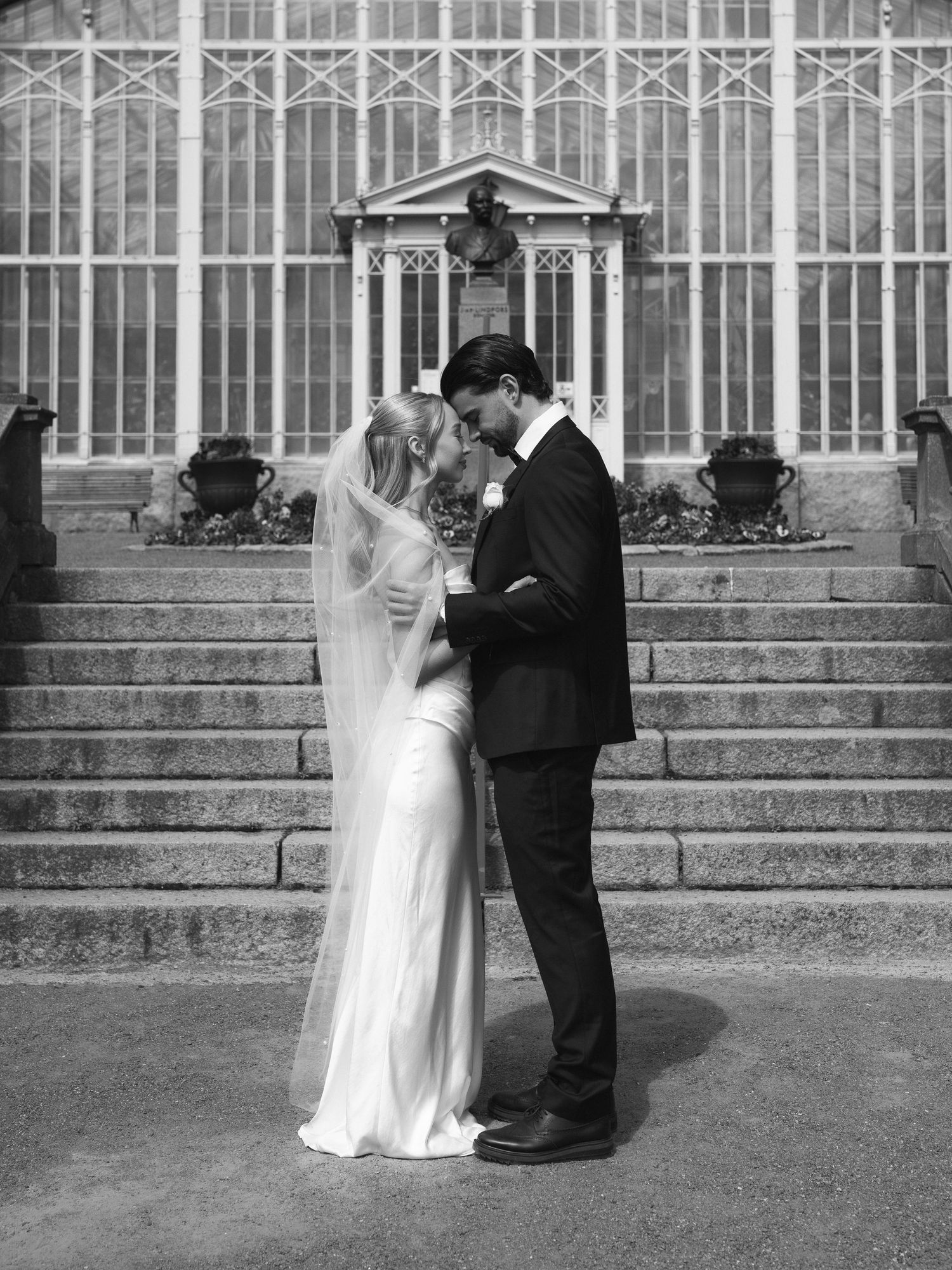 Black and white photo of a bride and groom sharing an intimate moment outside a grand greenhouse venue.