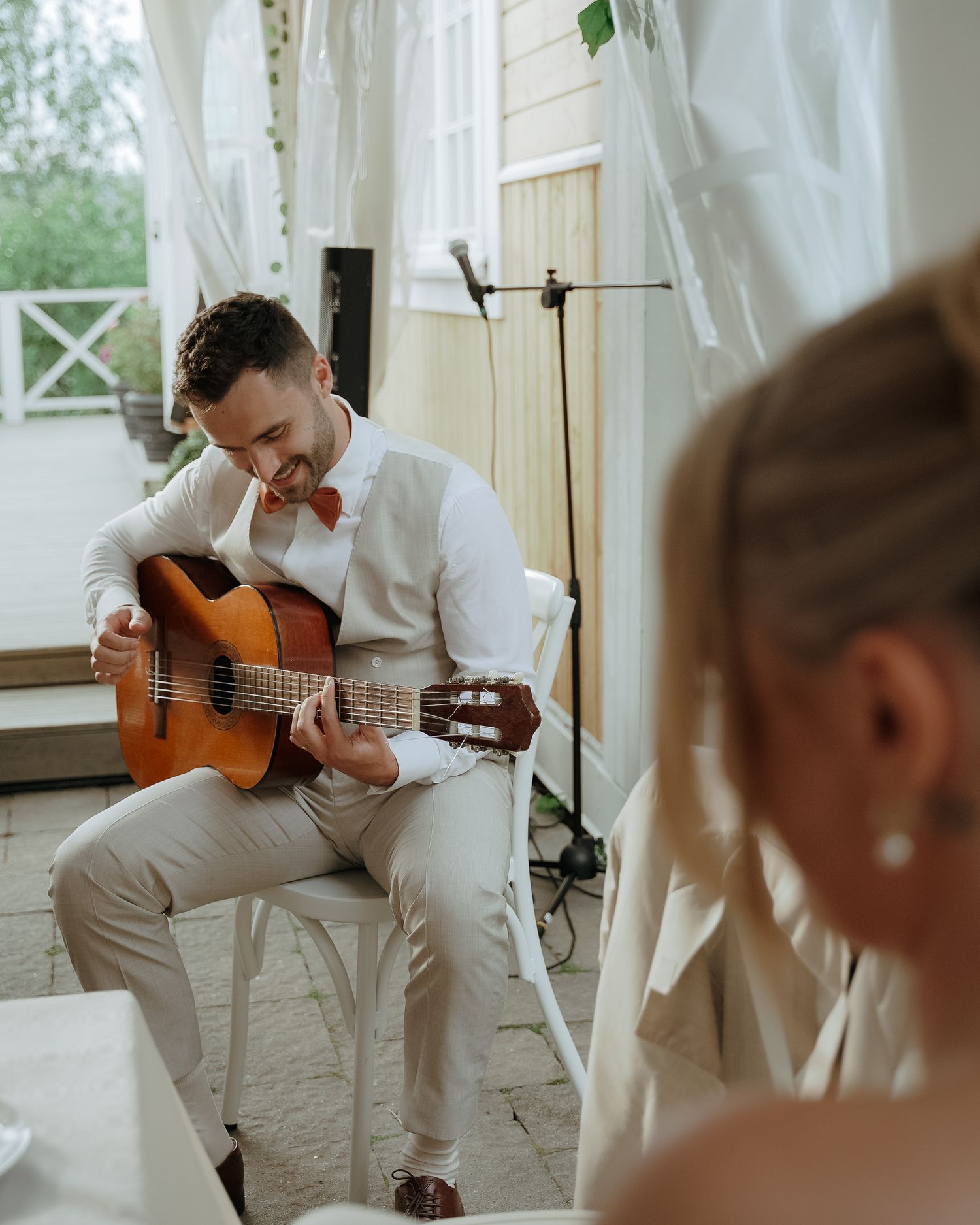 Musician in white attire performs on acoustic guitar by window with flowing curtains during intimate indoor performance.