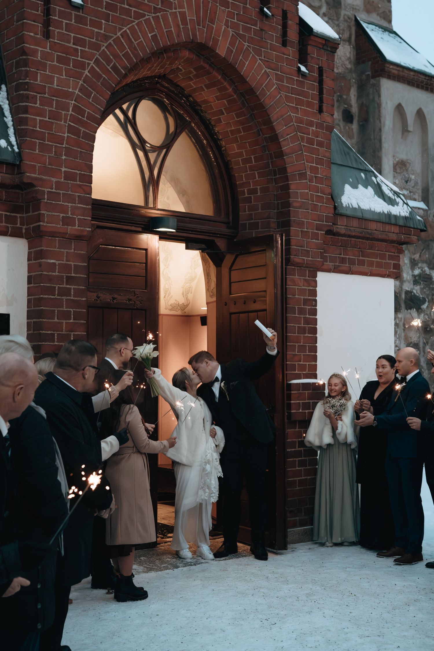 Guests gather outside a snowy church entrance with arched red brick doorway during a winter wedding celebration.