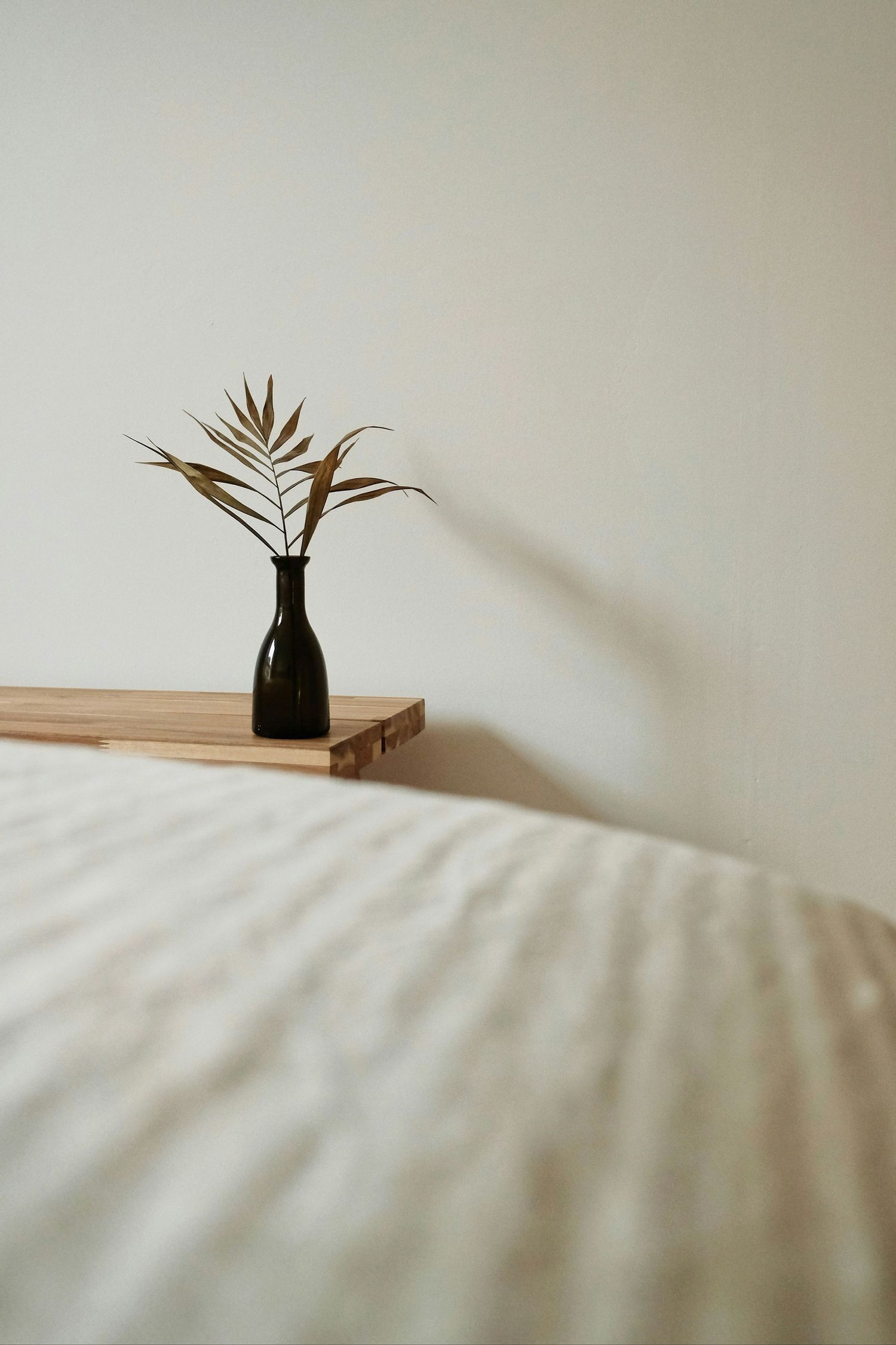 Minimalist black vase with palm leaf sits on wooden surface beside white bedding in soft natural light.