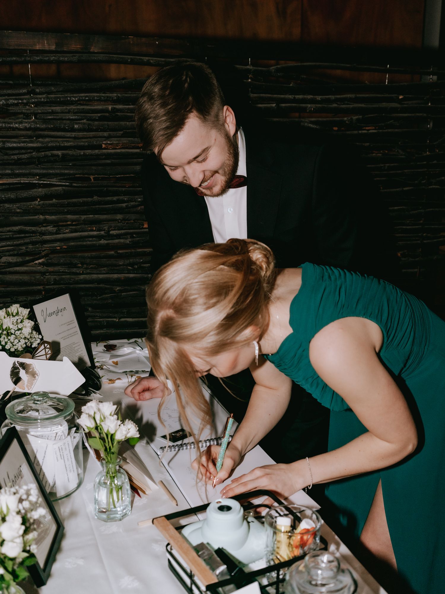 A couple at a rustic wedding reception sign their guest book at a table decorated with white flowers and mason jars.