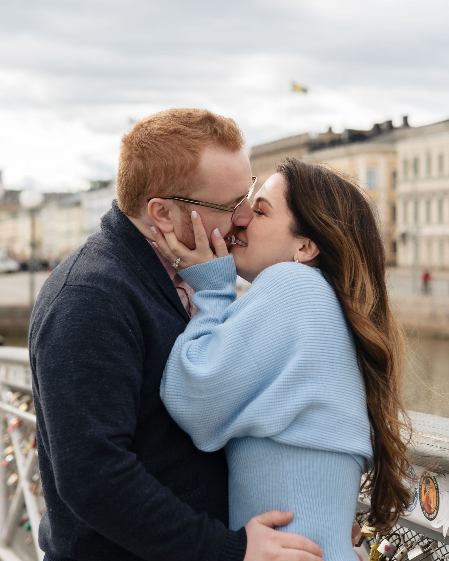 Couple sharing a romantic kiss on a bridge with love locks, woman in light blue sweater, man with red hair, city backdrop.