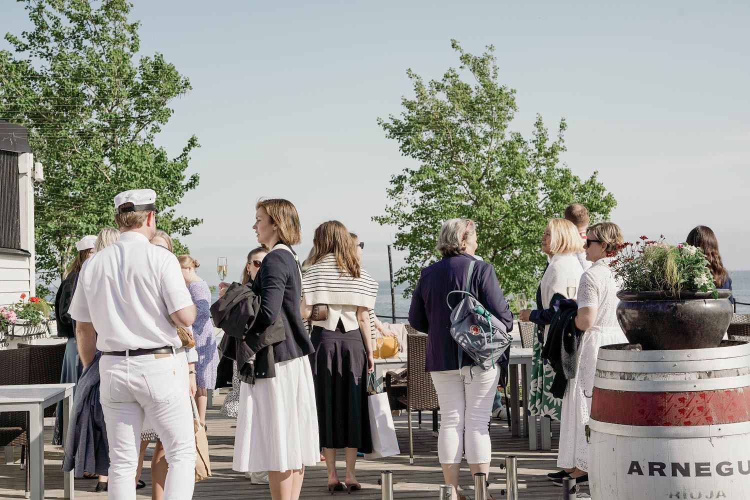 Guests gather on a sunny outdoor terrace near Arnegus barrels and lush green trees at a social event.