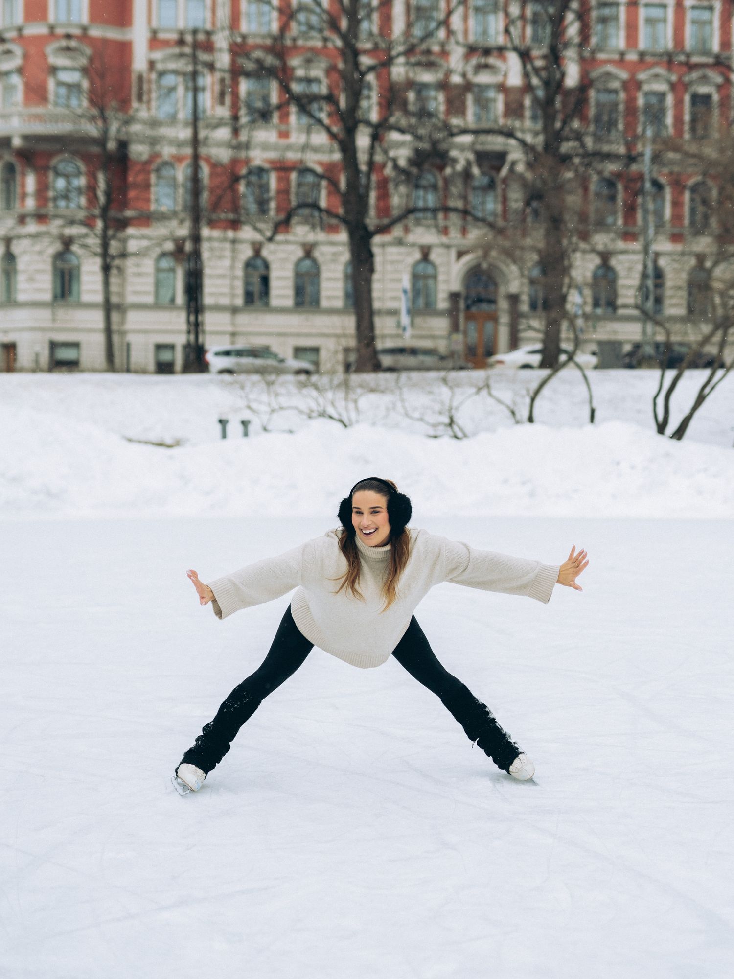 Figure in a white sweater and dark pants strikes a playful pose while ice skating in a snowy urban setting.