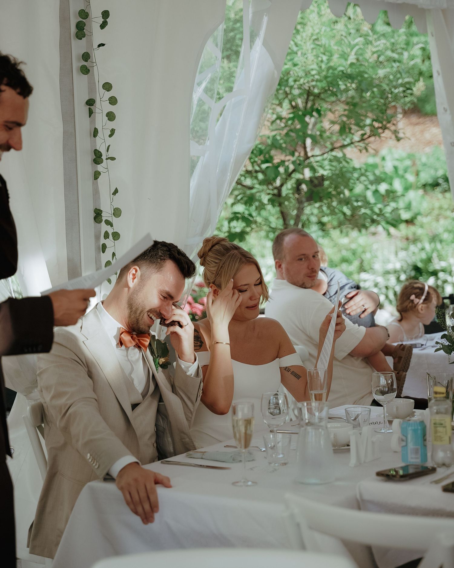 Wedding couple reacts emotionally during reception speeches while seated at white-clothed table under floral decor.