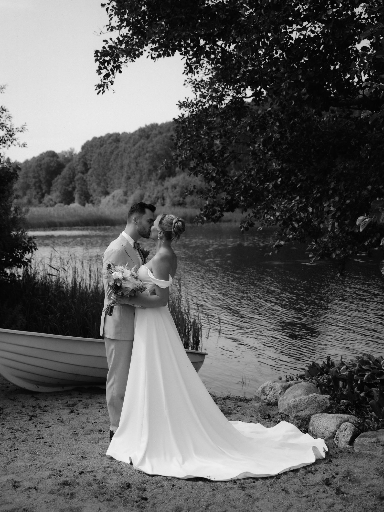 A couple in wedding attire embrace by a scenic lake with long flowing white dress trailing behind them.