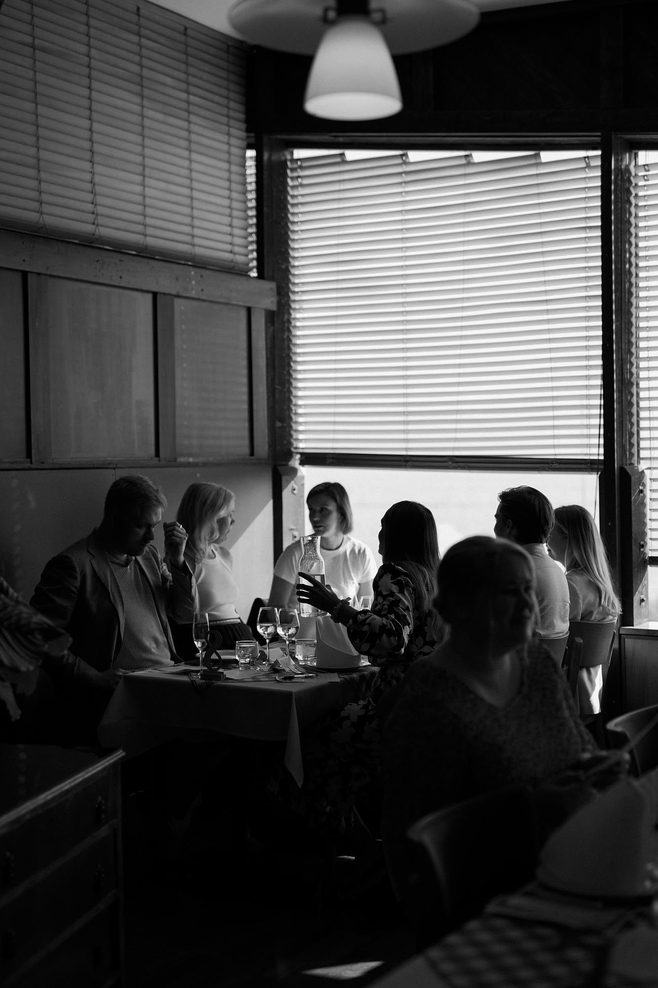 Black and white photo of a group dining in a cozy restaurant booth near window blinds with wine glasses on table.