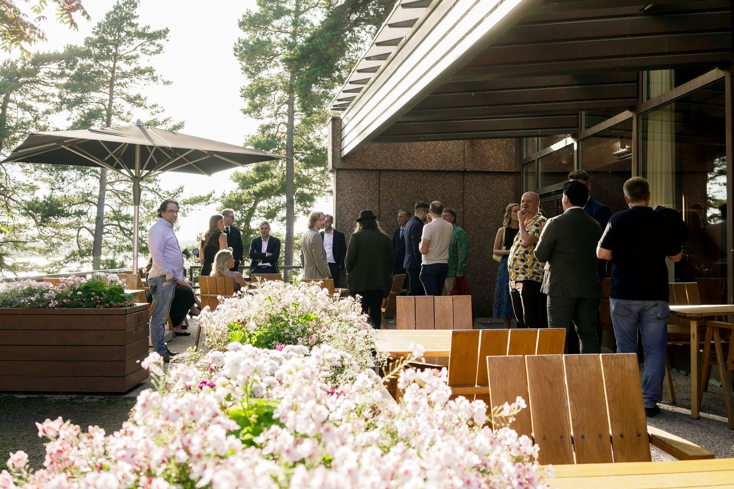 Guests mingle on a modern outdoor terrace surrounded by blooming flowers and pine trees at an elegant venue.