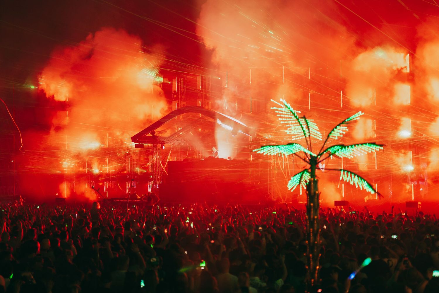 A neon palm tree glows against a red-lit stage at a concert with a large crowd silhouetted in the foreground.