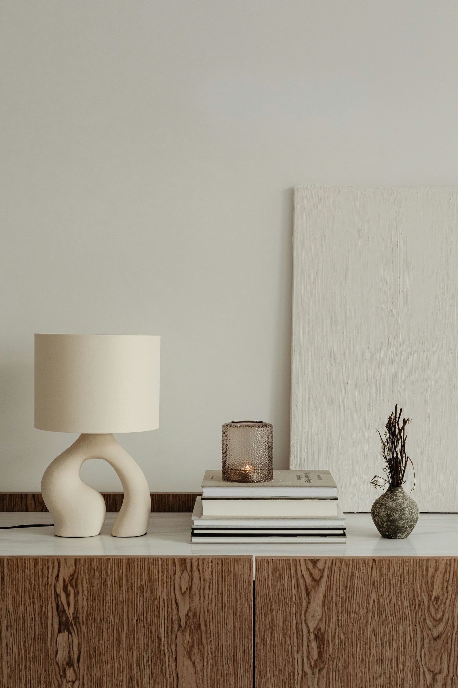 Modern minimalist decor display on wooden sideboard featuring curved ceramic lamp, books, and glass vase.